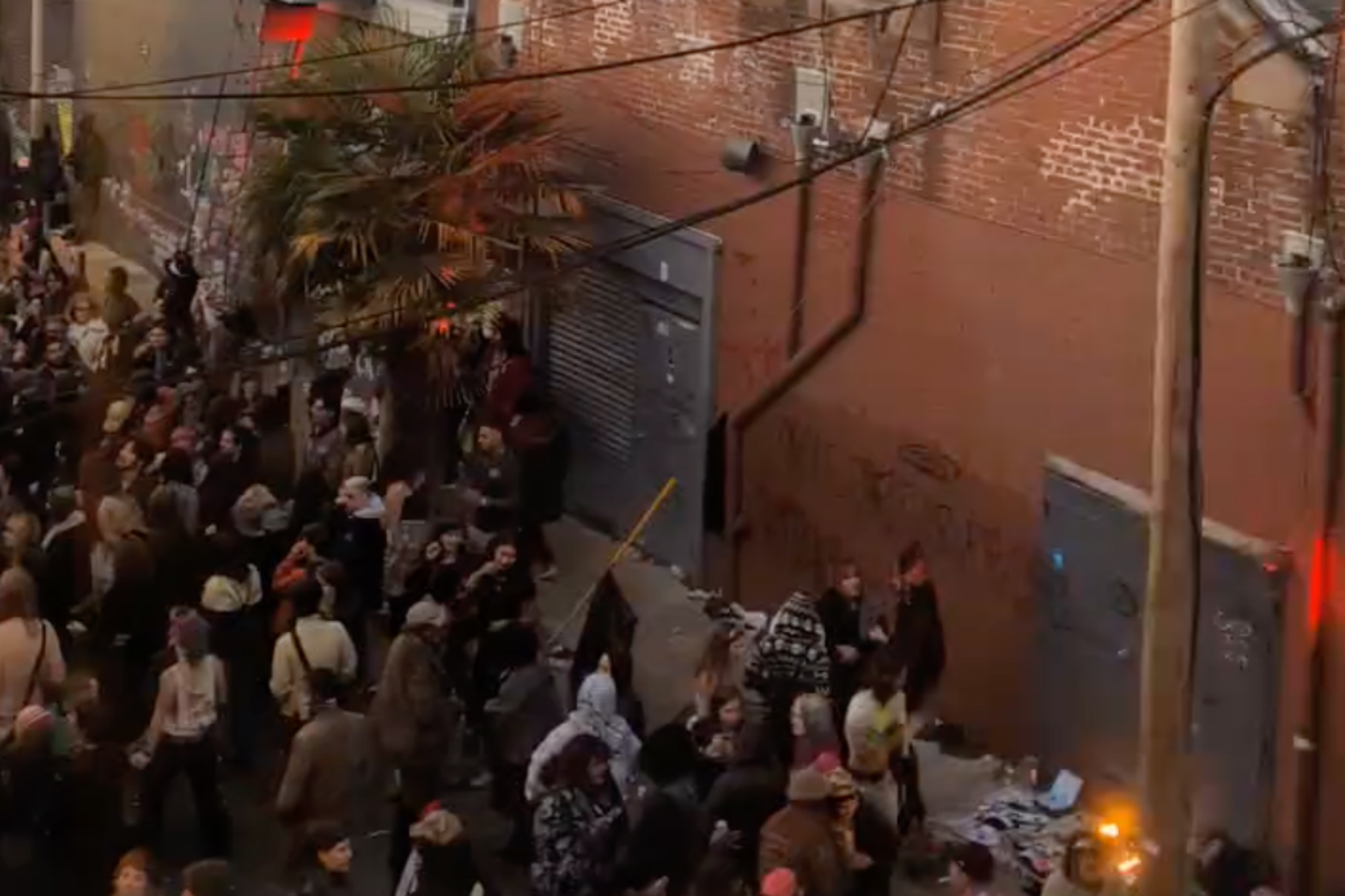 A crowd gathers on a street by a large building with a sign that reads "Free Palestine" painted on it. People stand on a balcony and a fire burns nearby.