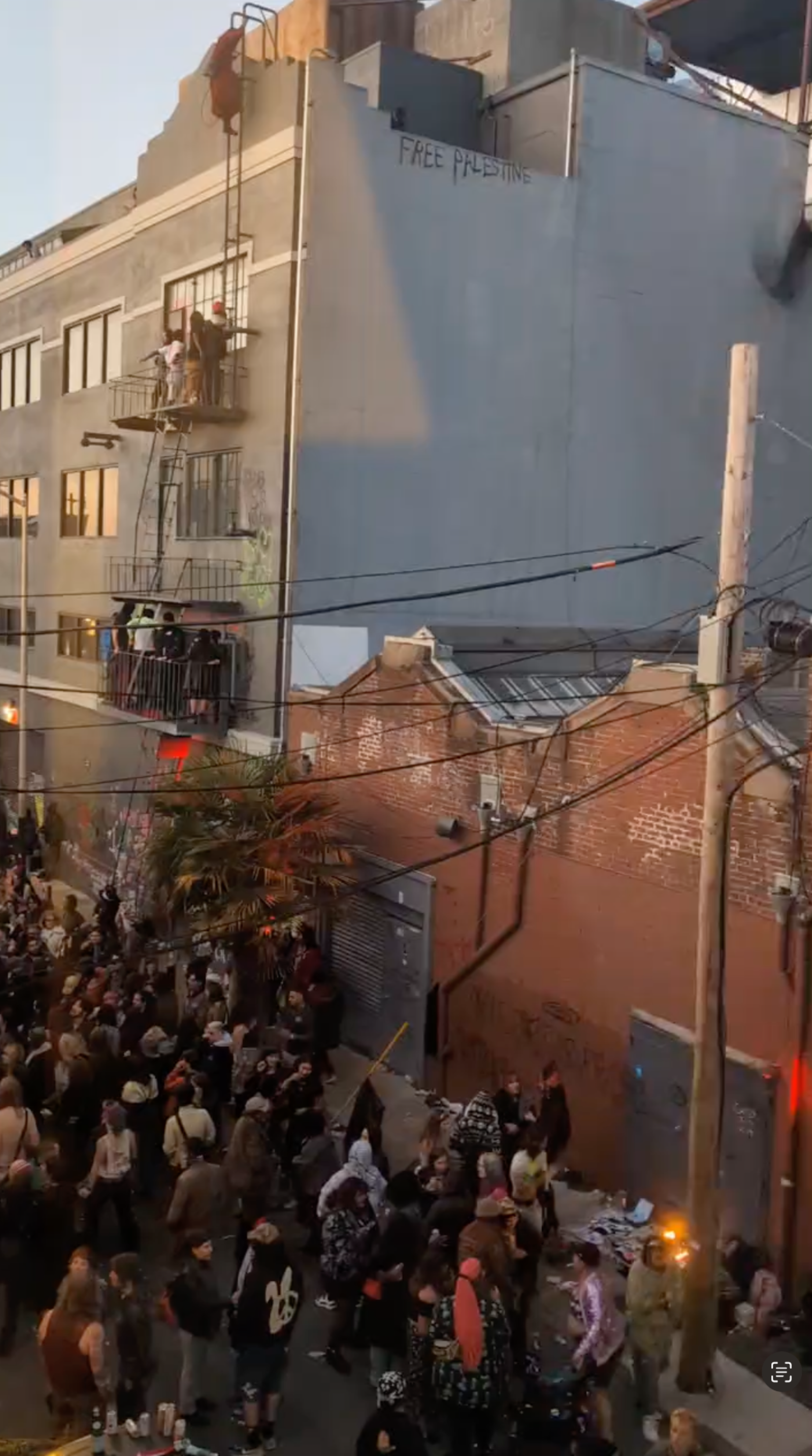 A crowd gathers on a street by a large building with a sign that reads "Free Palestine" painted on it. People stand on a balcony and a fire burns nearby.