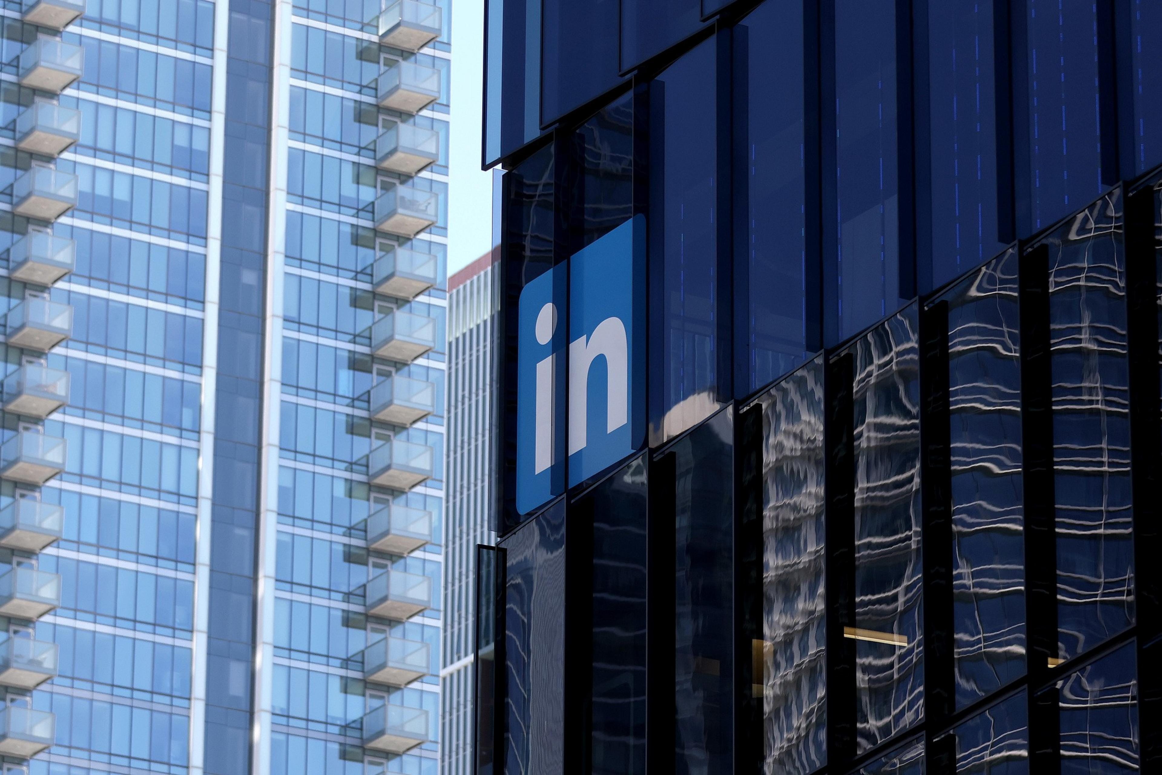 A large blue LinkedIn logo with white "in" letters is mounted on a modern glass office building, with adjacent high-rise buildings reflecting on its surface.