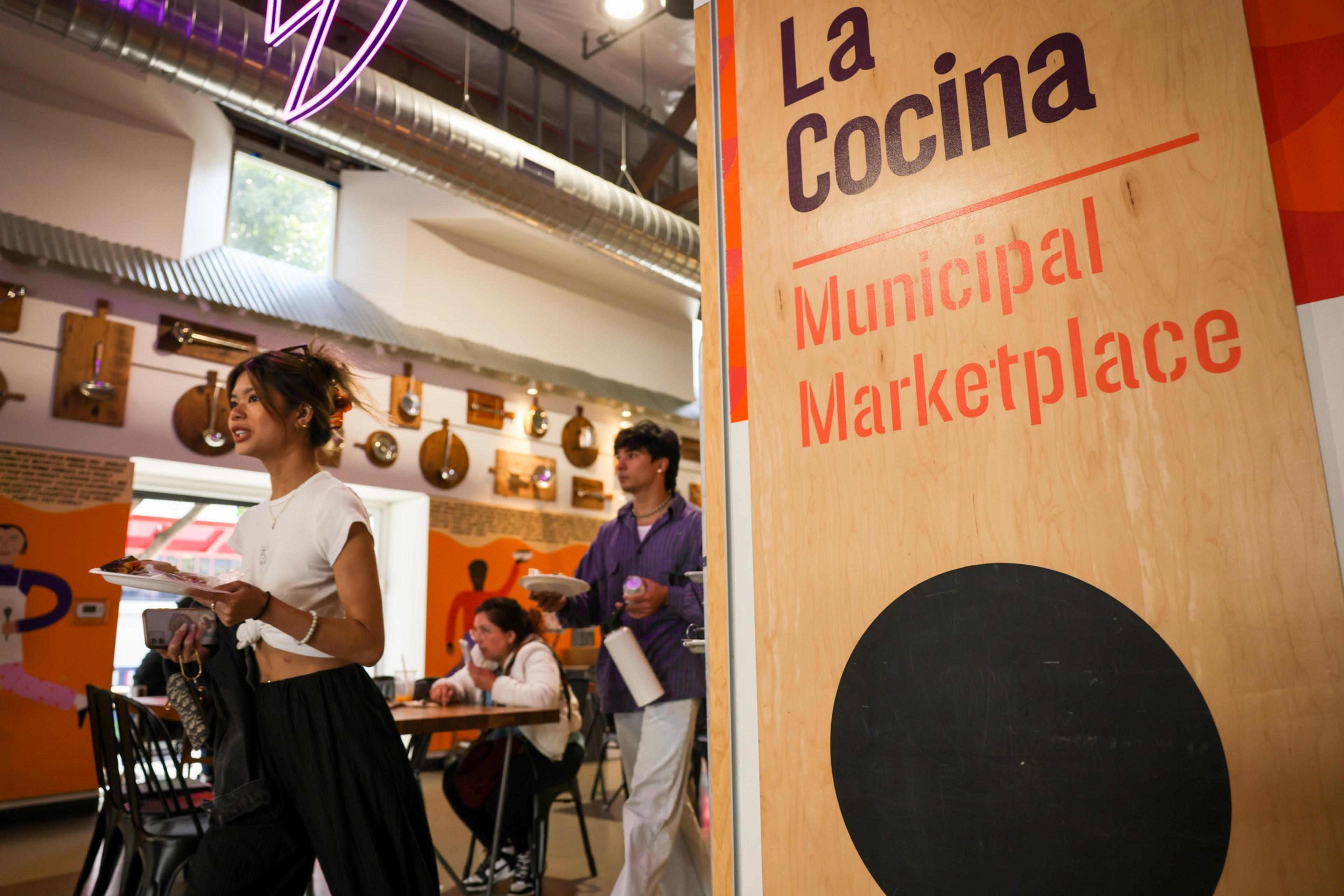 A bustling marketplace with people carrying plates, a neon sign above, and a sign that reads "La Cocina Municipal Marketplace."