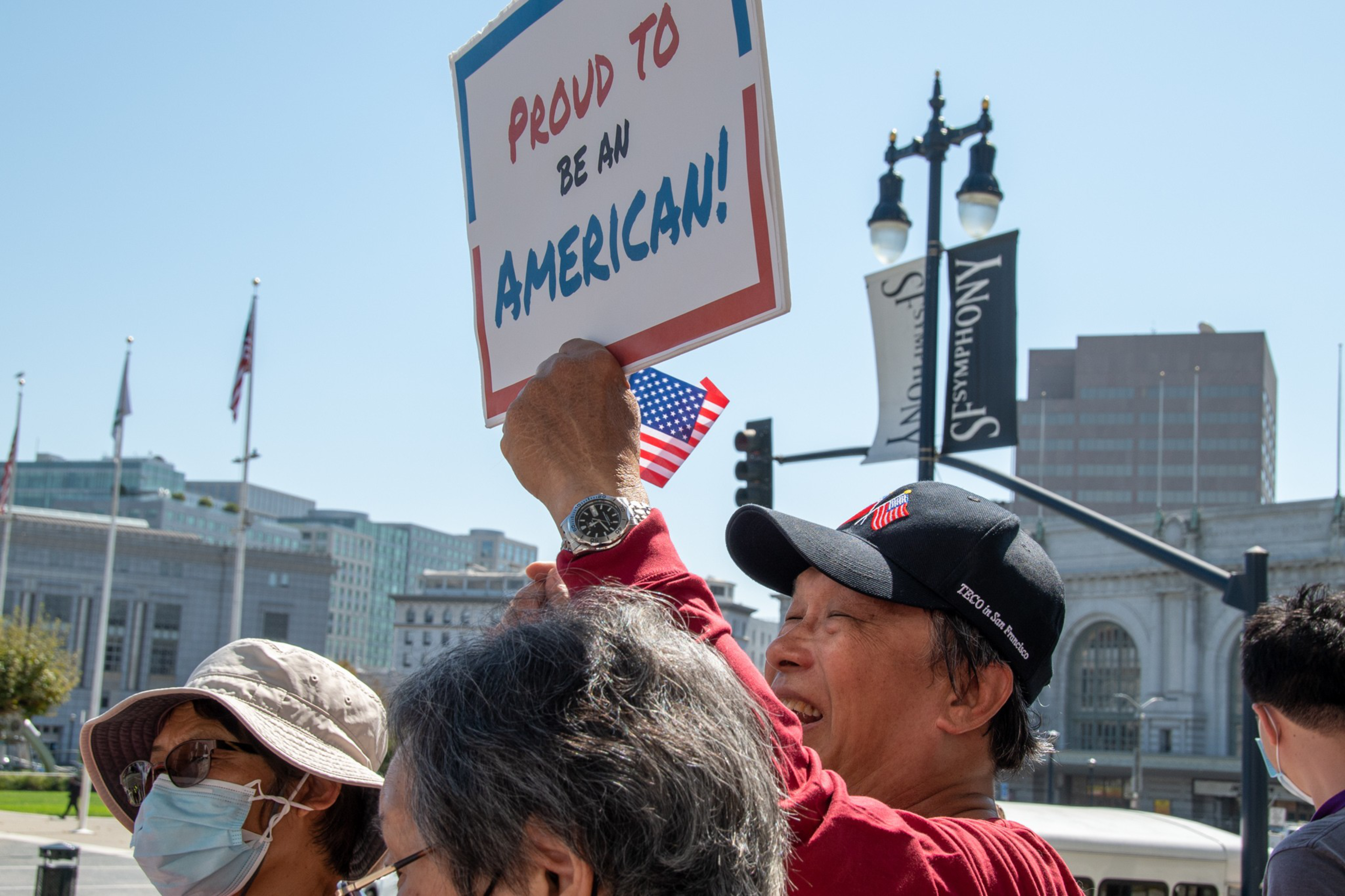 A person wearing a black cap holds a sign saying "Proud to be an American!" while others around wear hats and face masks outdoors in a city.