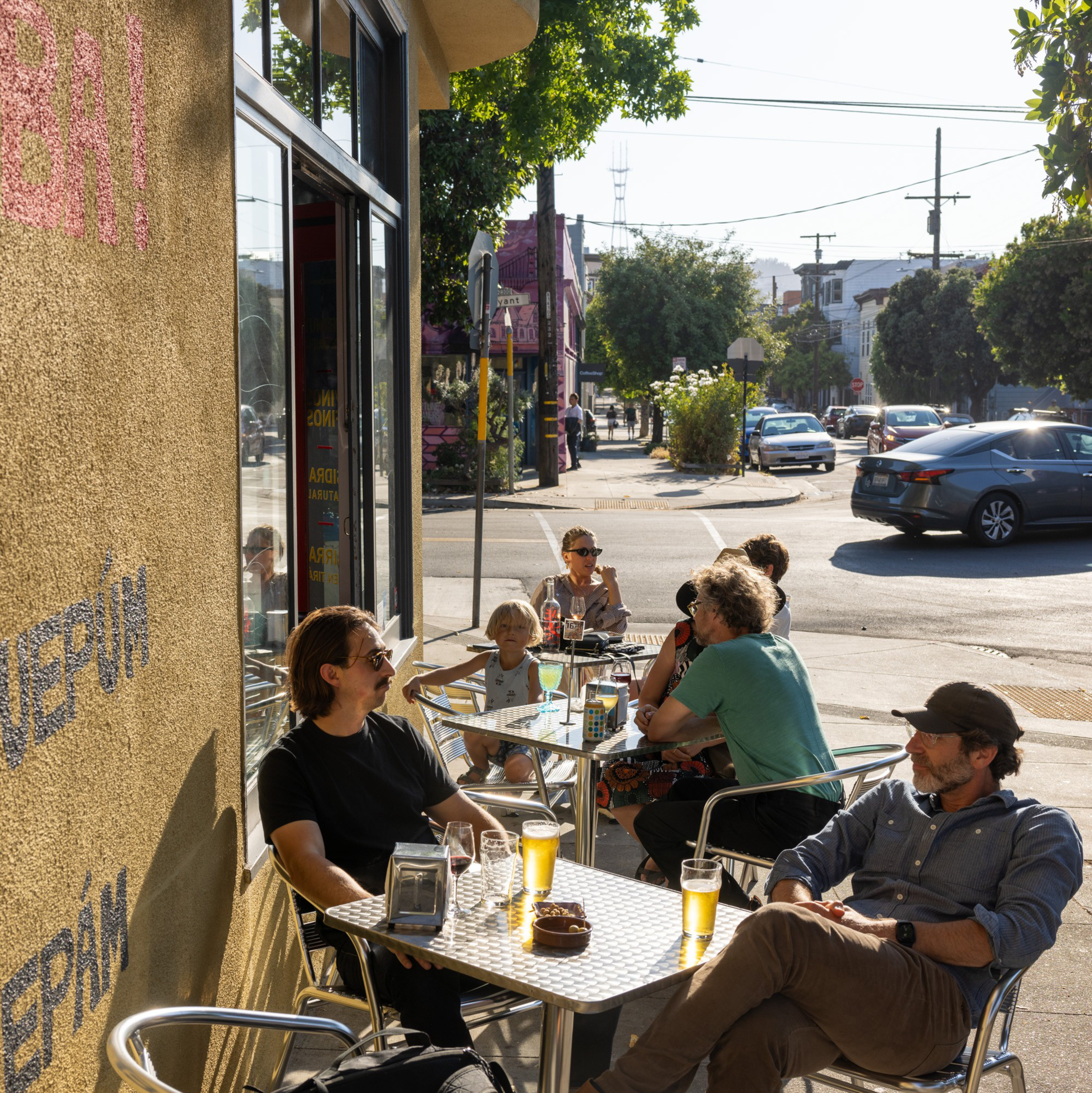 People are sitting at outdoor tables of a café on a sunny day, enjoying drinks and casual conversation. The café is on a street corner with cars and trees nearby.