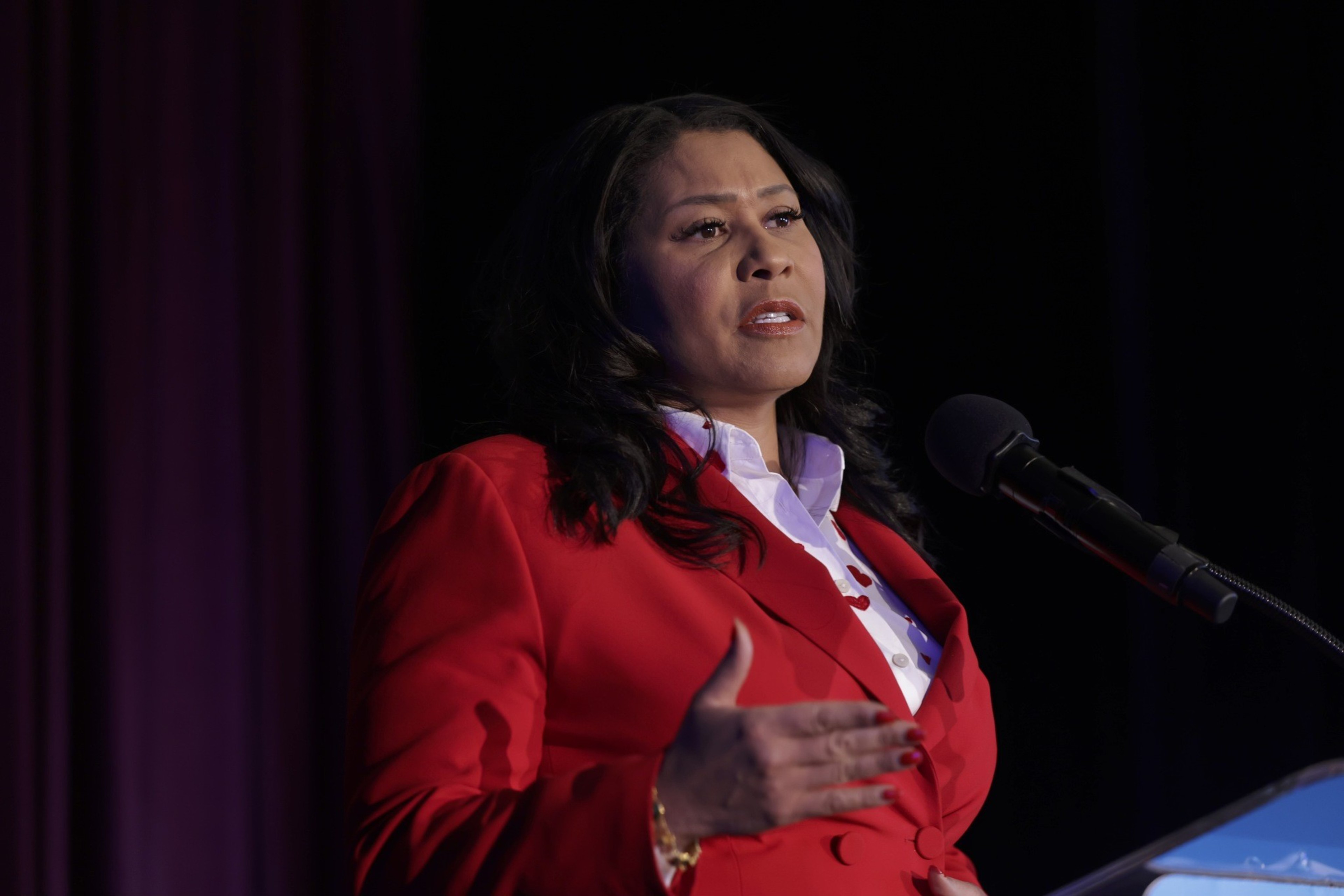 A woman in a red blazer speaks at a podium with a microphone, against a purple backdrop.
