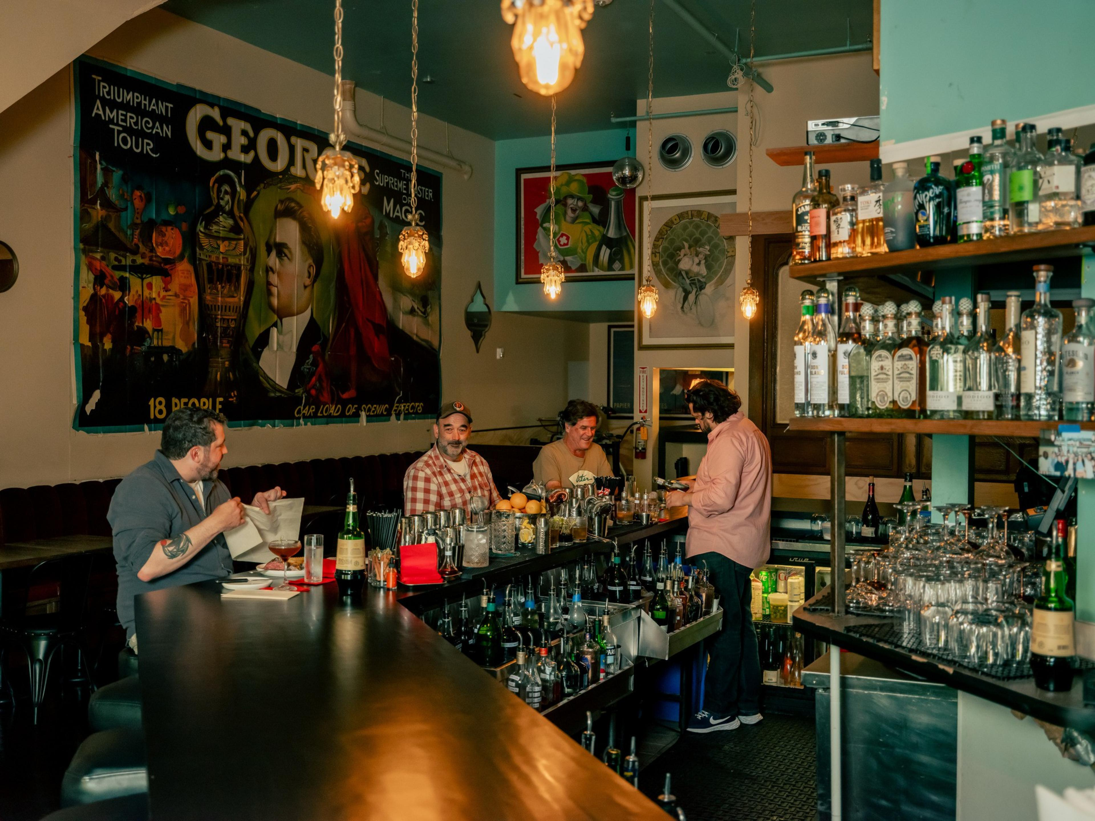 A bar scene with three men interacting. Shelves of assorted liquor bottles are in view. A vintage magic show poster decorates the wall. Warm lighting adds ambiance.