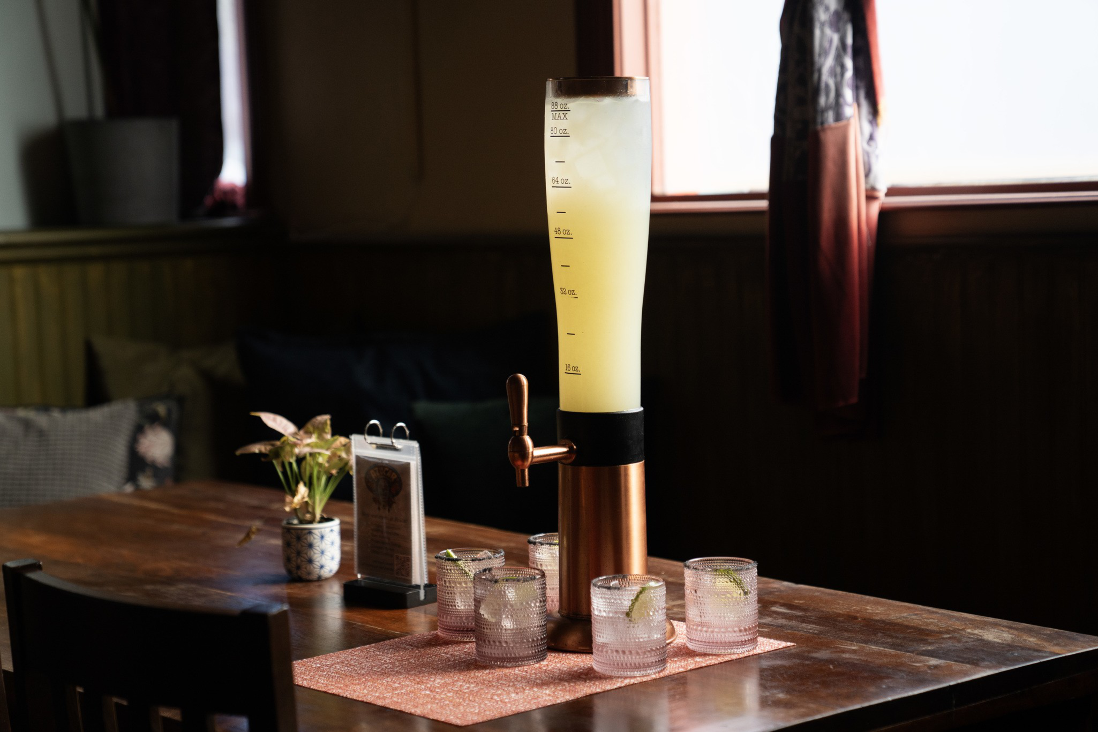 A tall drink dispenser with yellow beverage and four glasses with ice and lime wedges sit on a wooden table with a pink placemat in a dimly lit room.