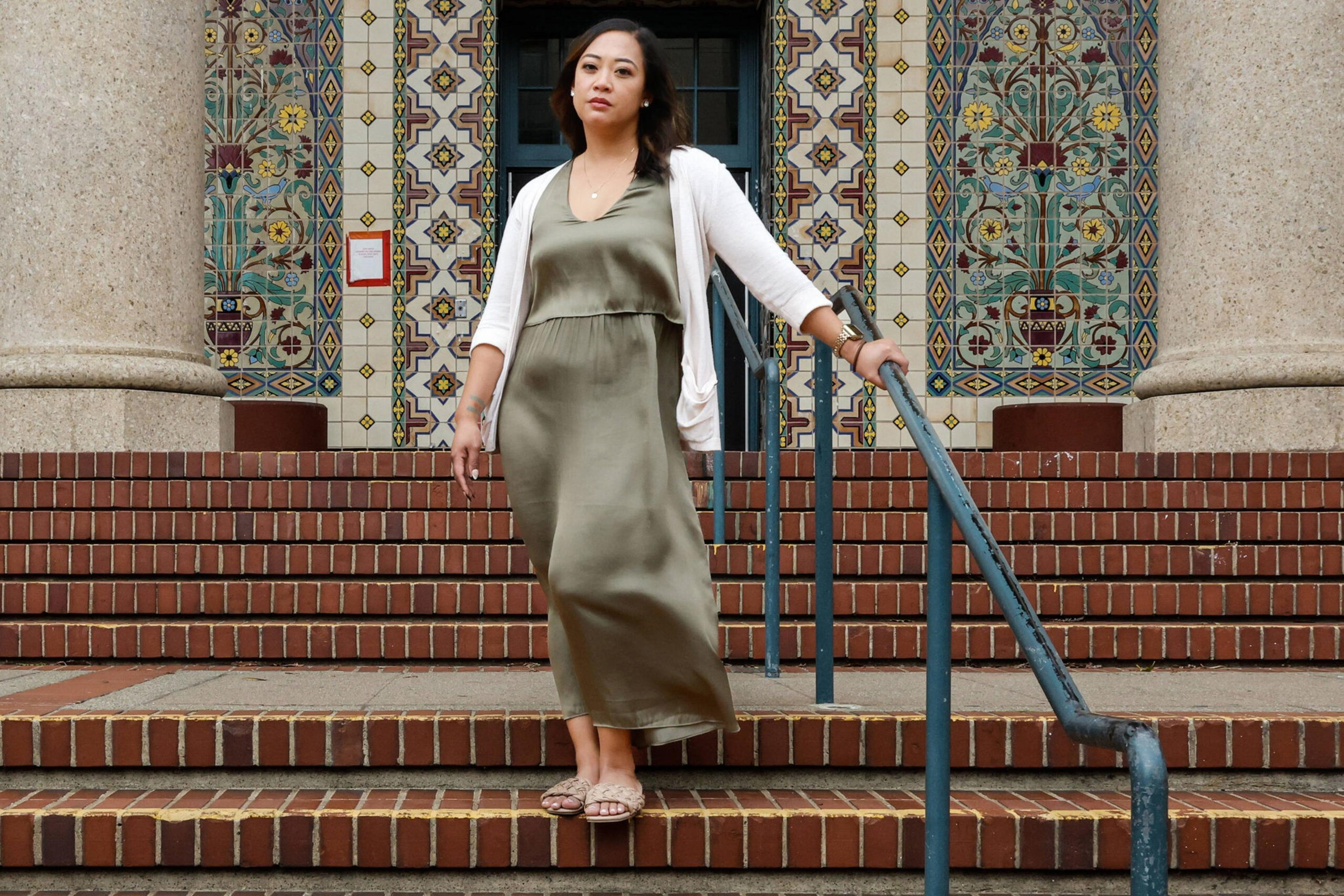 A woman in a green dress and white cardigan stands on brick steps, holding a metal railing, with an ornate tiled wall and columns behind her.