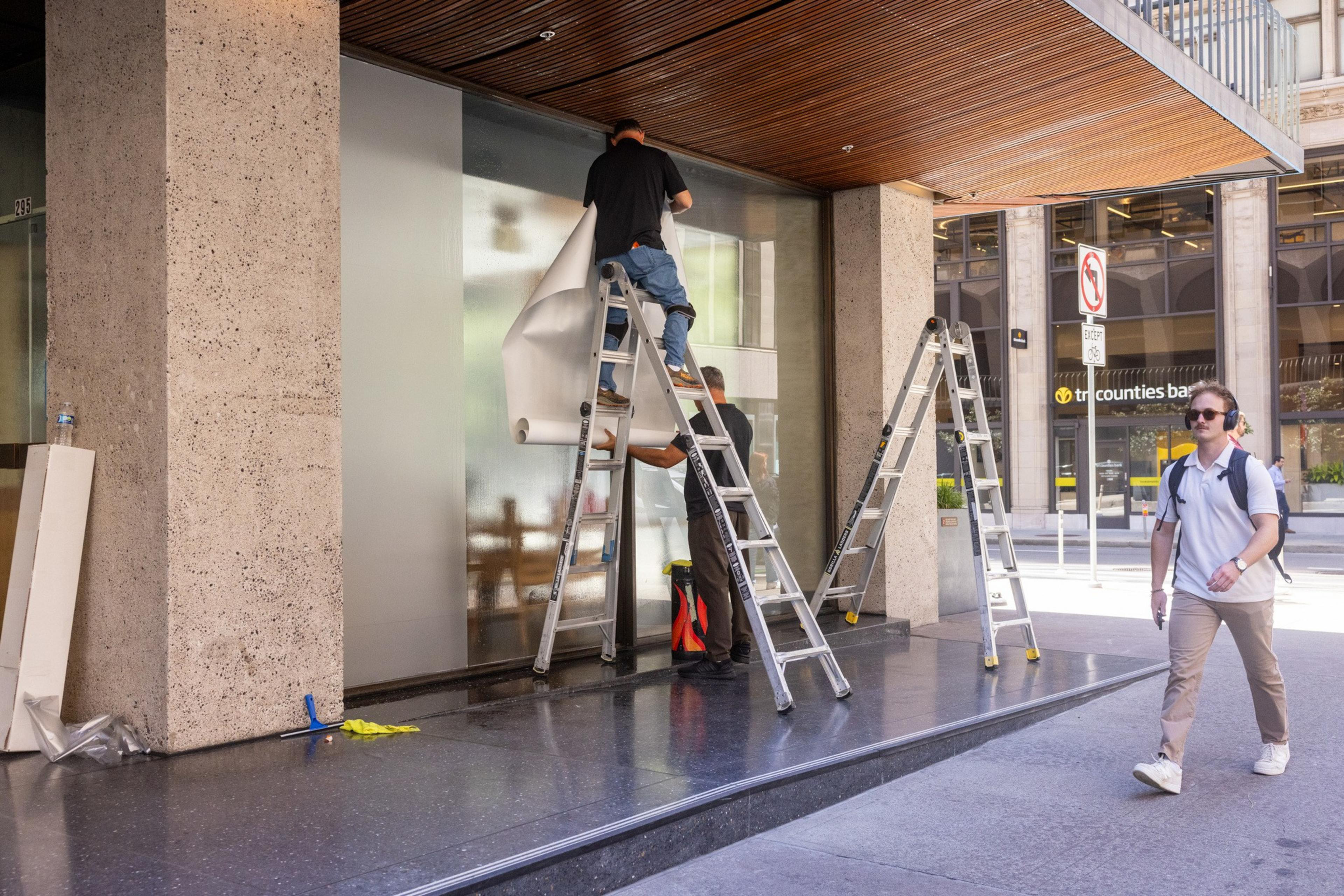 A man on a ladder is removing or applying a large sheet to a glass storefront while another man assists below; a pedestrian walks by on the sidewalk.