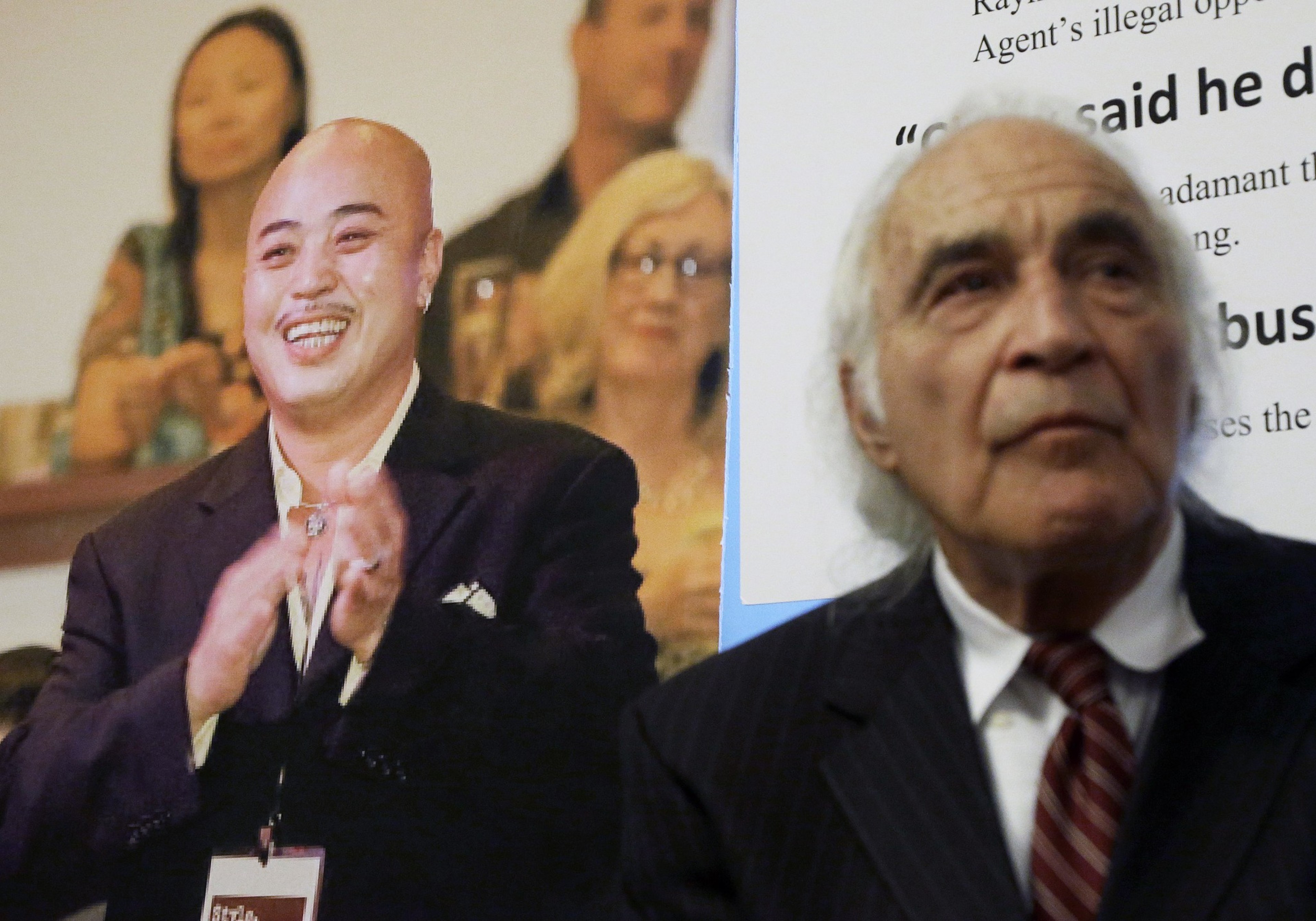 A man, who is out of focus, looking into the distance with a serious expression with a photo behind him of a smiling man.