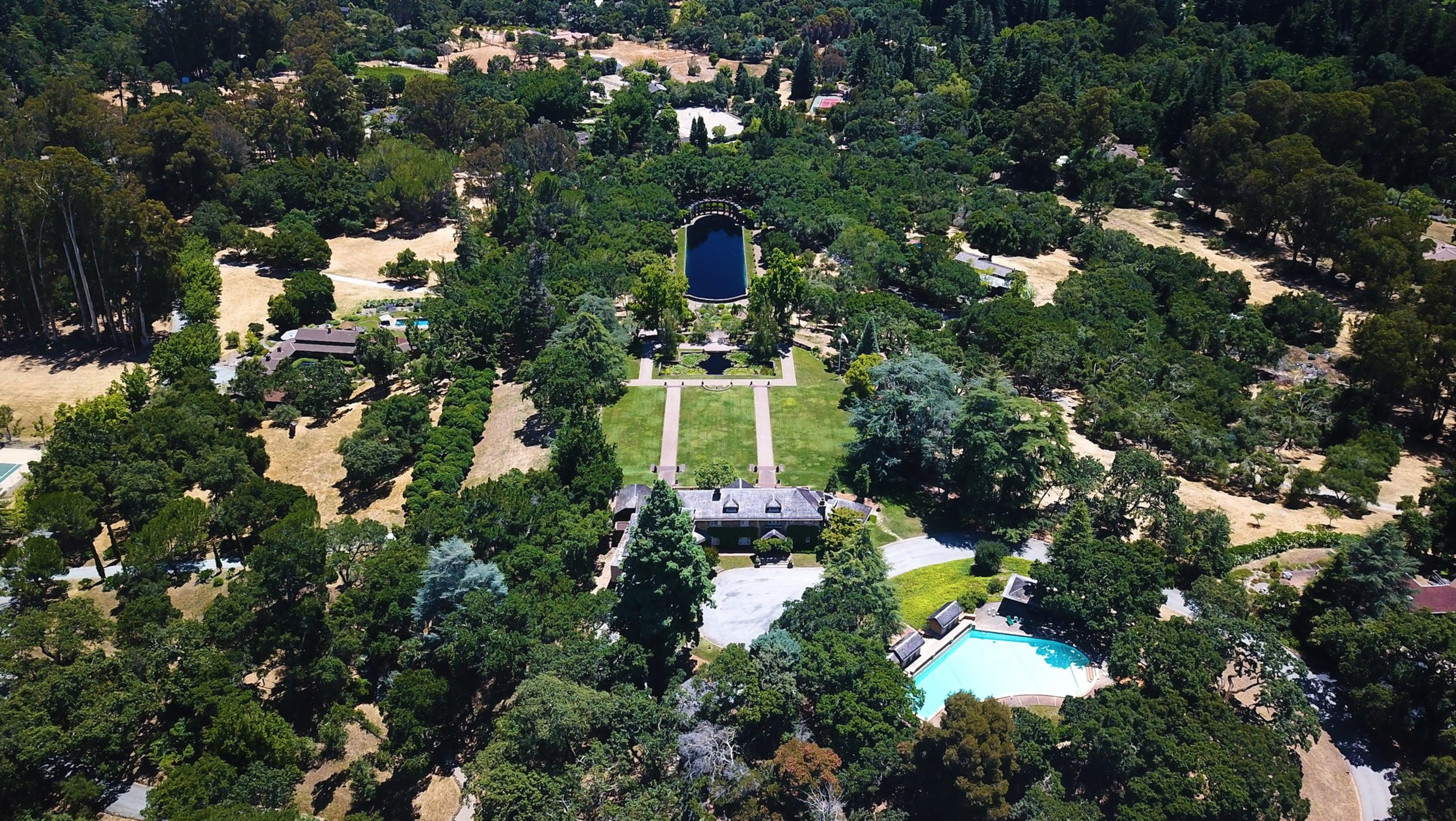 A large estate features a central house, a rectangular lawn, a rectangular pond beyond it, and a swimming pool surrounded by dense trees.