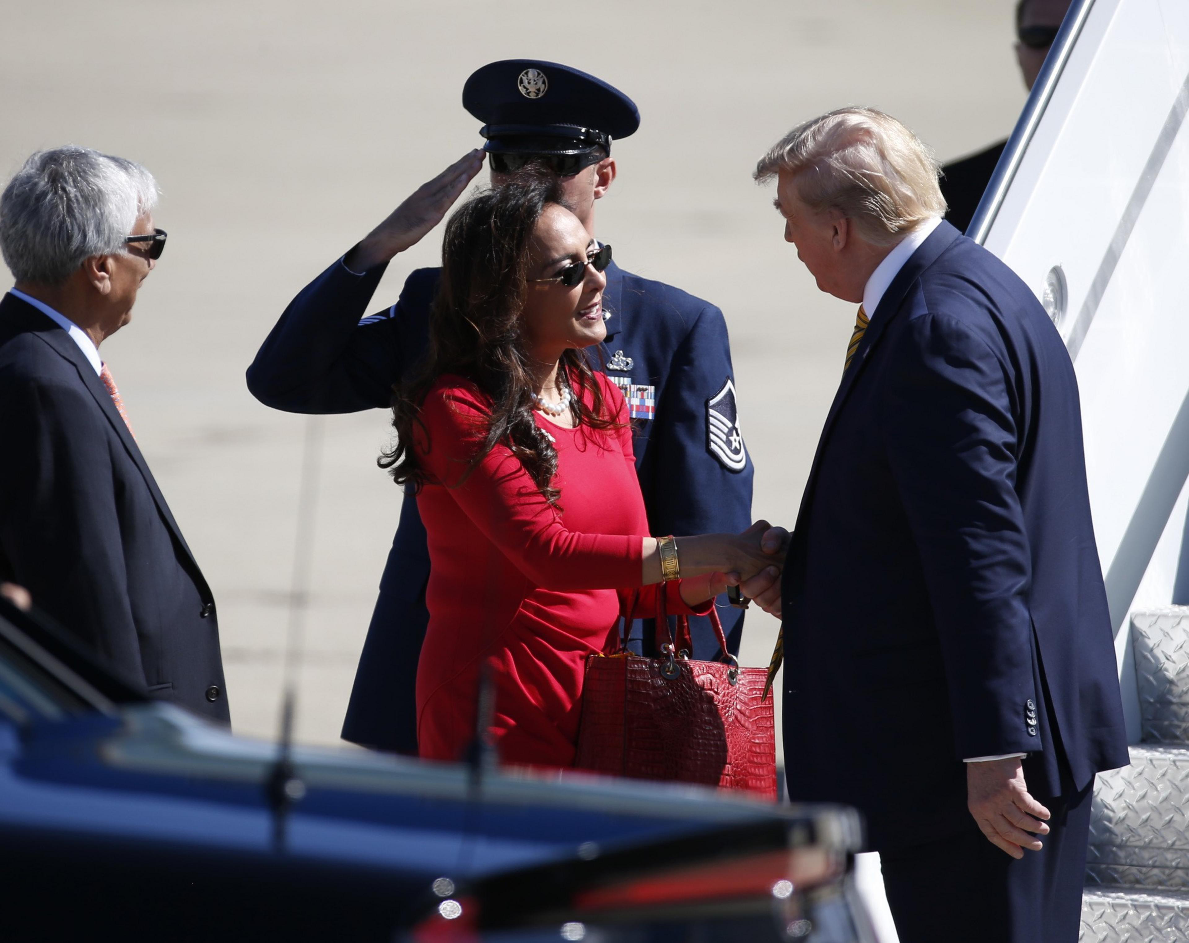 A woman in red shakes hands with a man in a suit near an airplane staircase. A man in a military uniform salutes, and another person stands nearby.