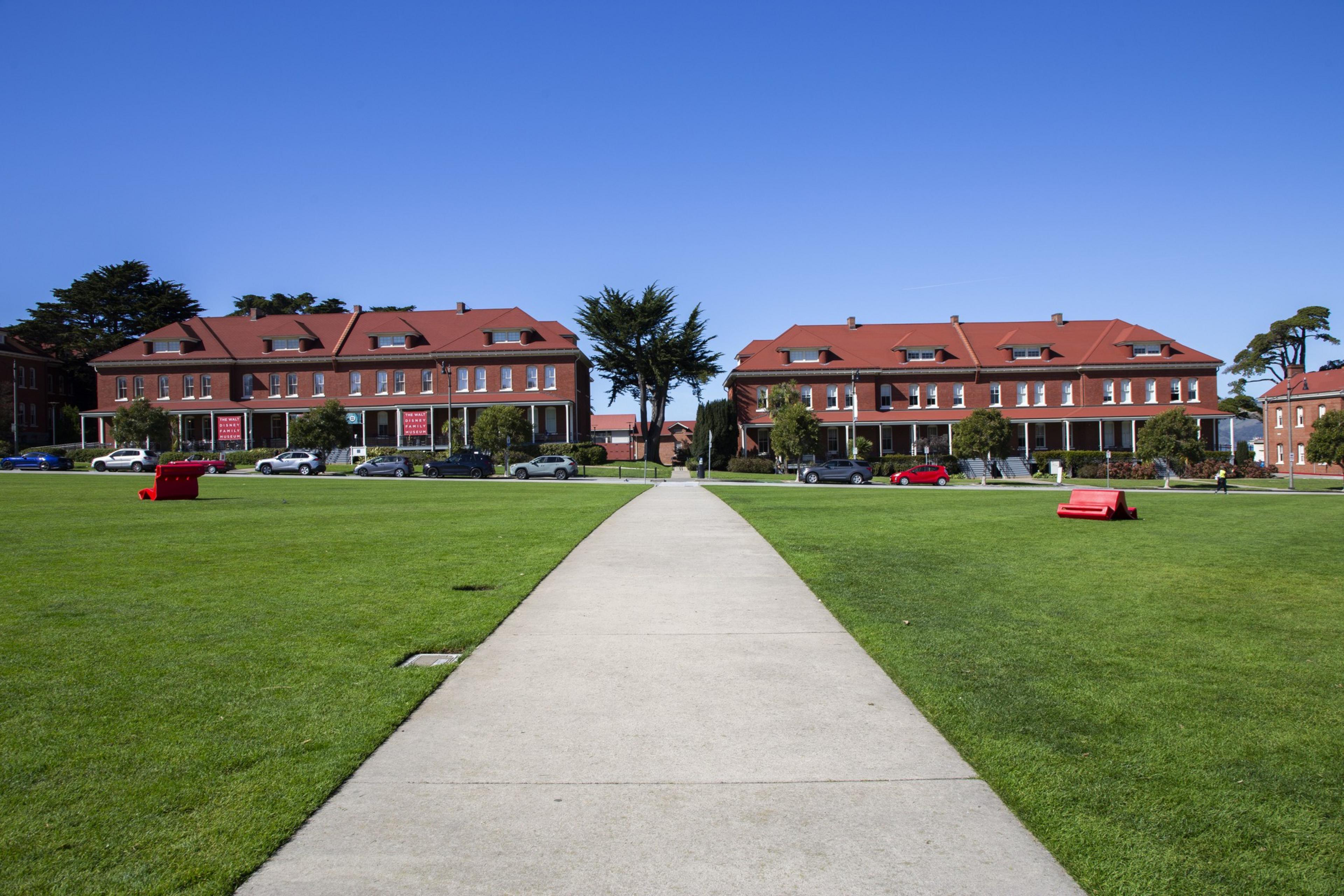 A path leads through a grassy field towards two large, identical brick buildings with red roofs. Trees and cars line the street, under a clear blue sky.