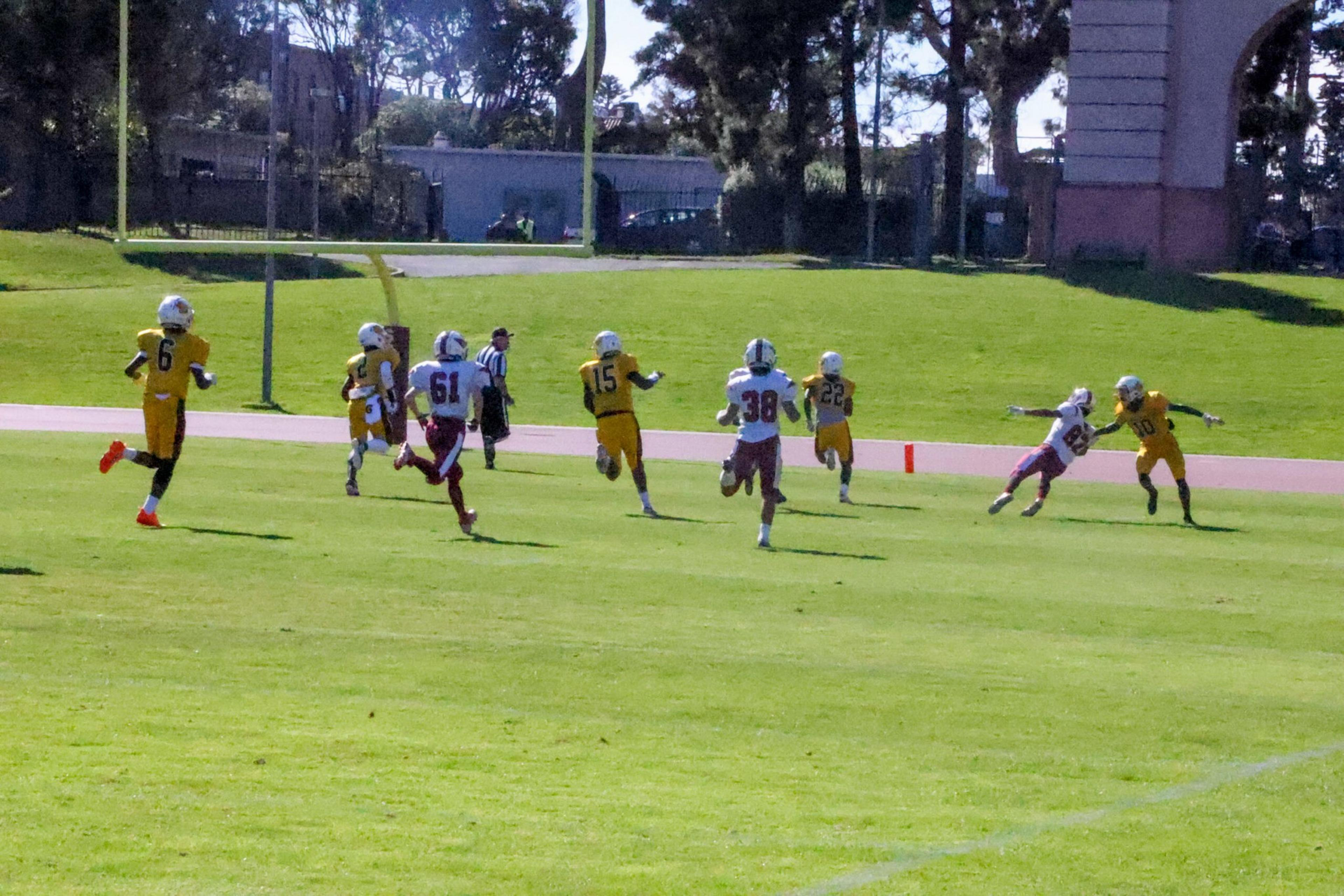 Football players in yellow and white uniforms run toward the end zone, with a player in yellow breaking a tackle near the goal line.