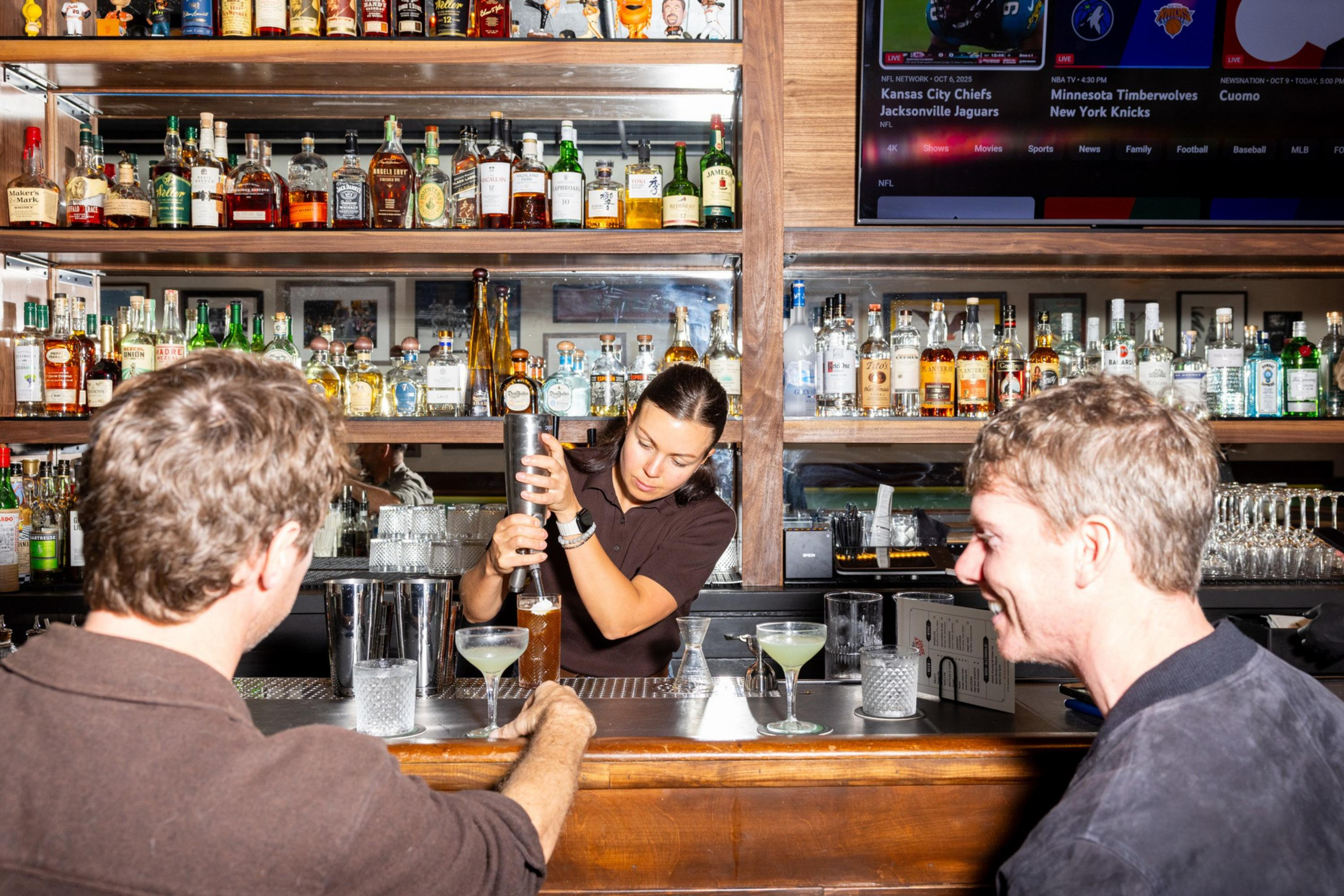 A bartender is pouring a drink while two men sit at the bar, smiling, with shelves of liquor bottles and a TV screen in the background.