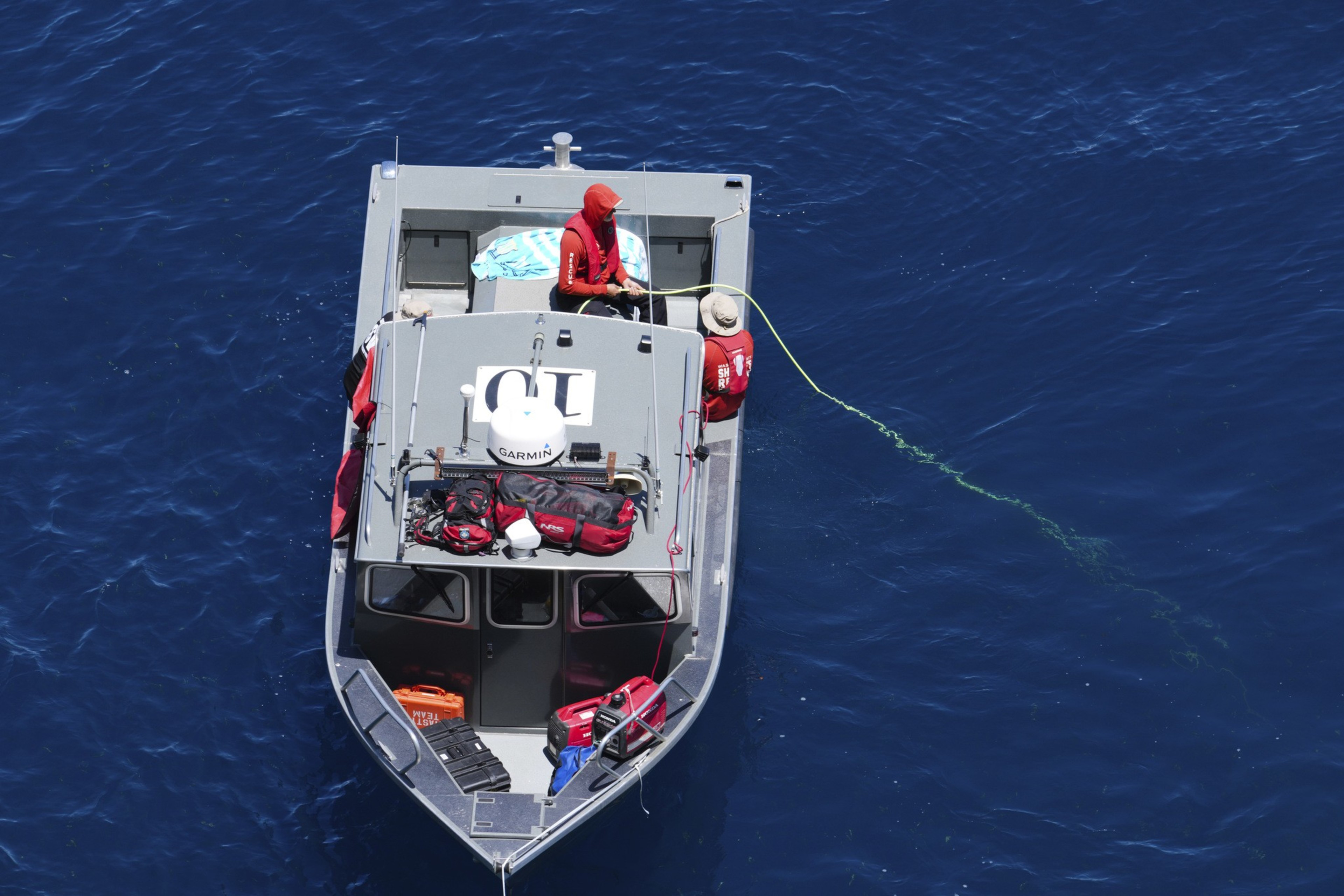 Two people in red clothing are on a small gray boat in deep blue water. One person holds a green rope extending into the water. Onto the boat's roof, various gear is secured.