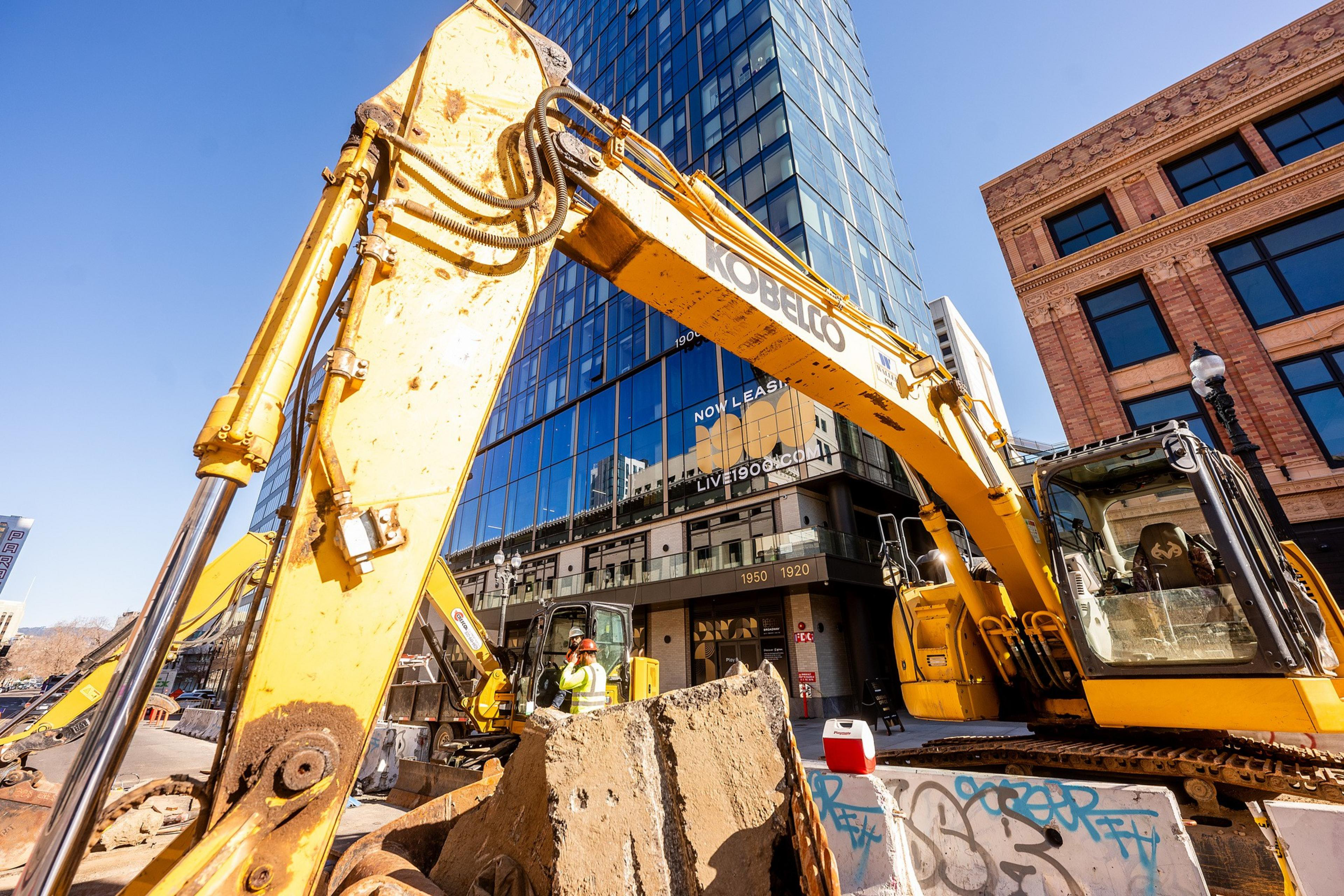 A yellow excavator operates near modern glass and old brick buildings, with a construction worker nearby. Graffiti covers a concrete barrier in the foreground.