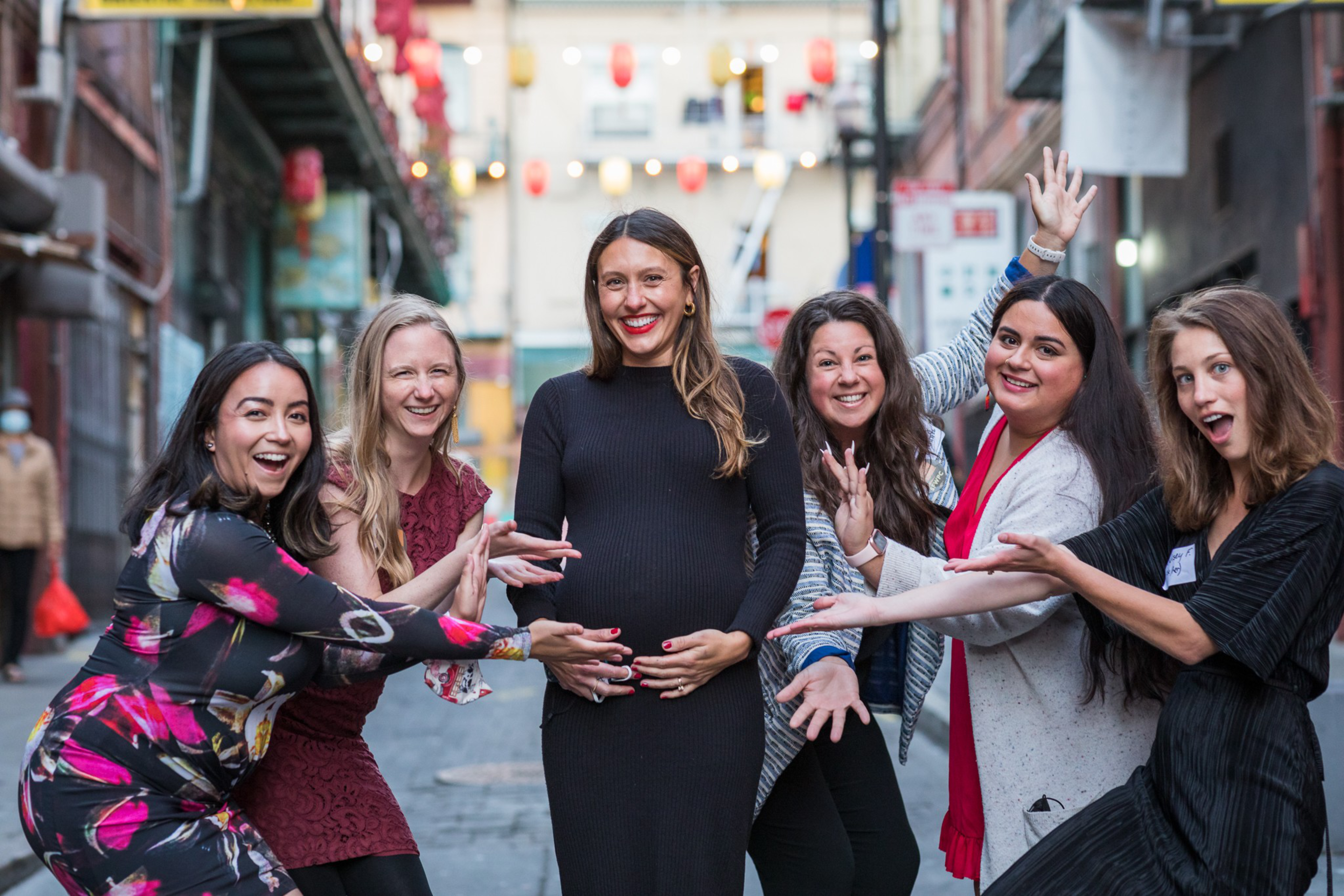 Six women stand in a row on a city street, smiling and gesturing excitedly toward the pregnant woman in the black dress at the center.