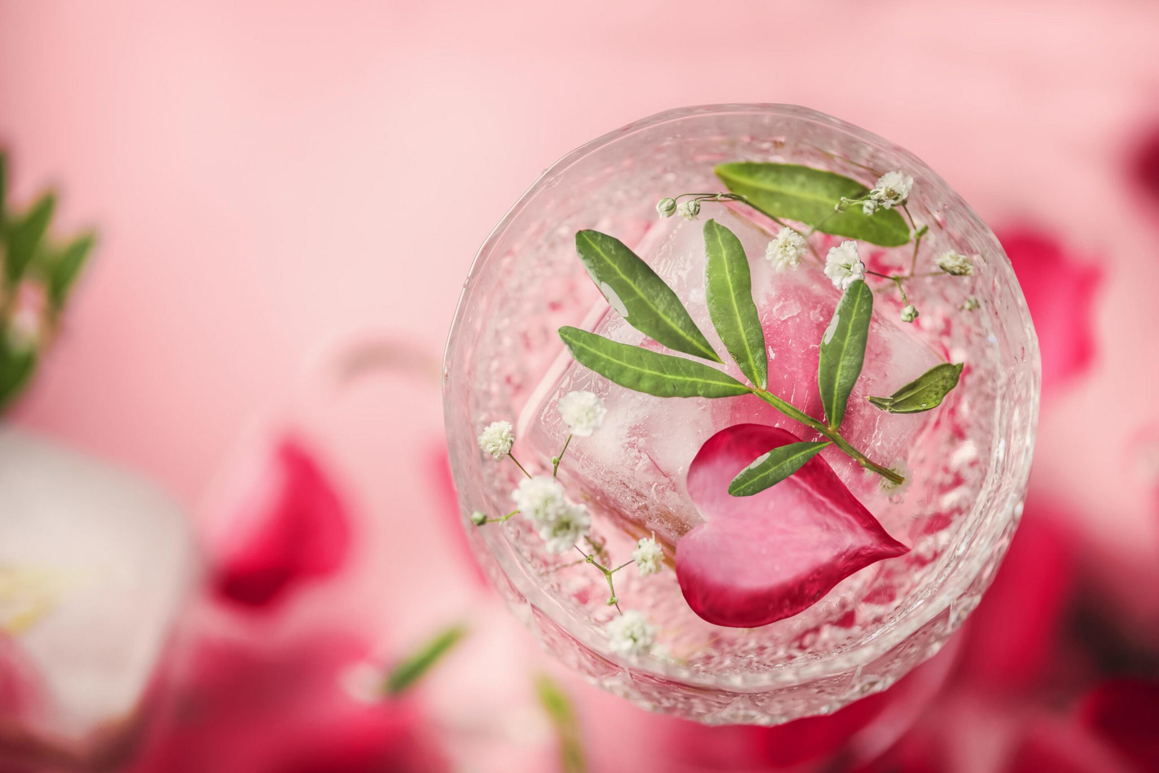 A glass filled with a clear beverage contains ice cubes, a heart-shaped pink petal, small white flowers, and green leaves, set against a pink background.