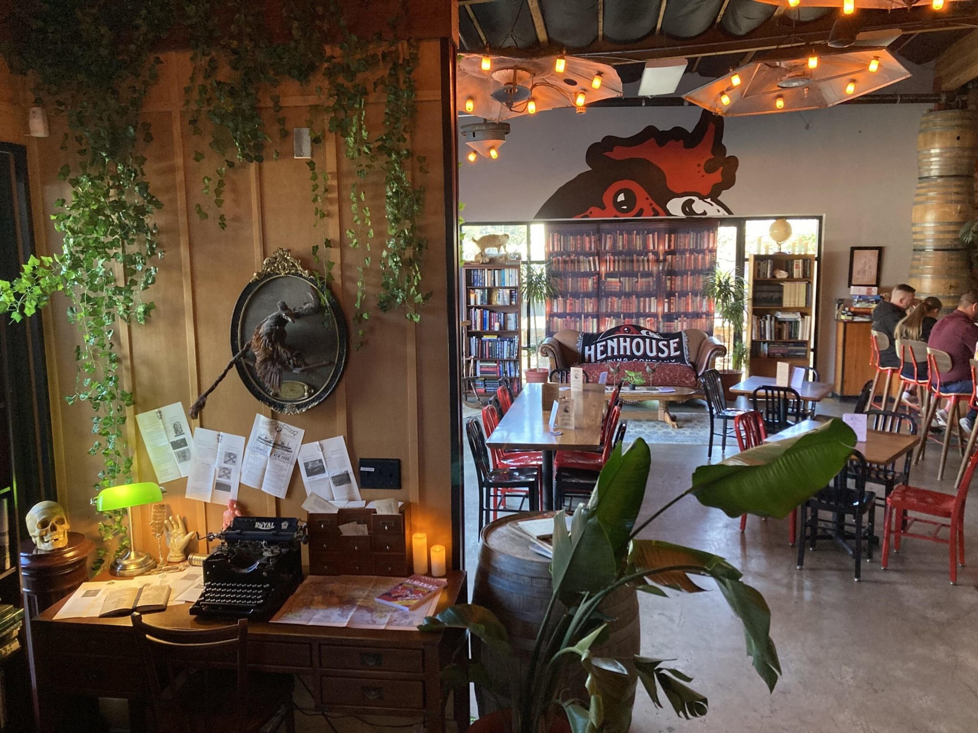 The image shows a cozy cafe with wooden furniture, bookshelves, and greenery. A vintage desk holds papers and a typewriter. A painted chicken mural decorates the wall.