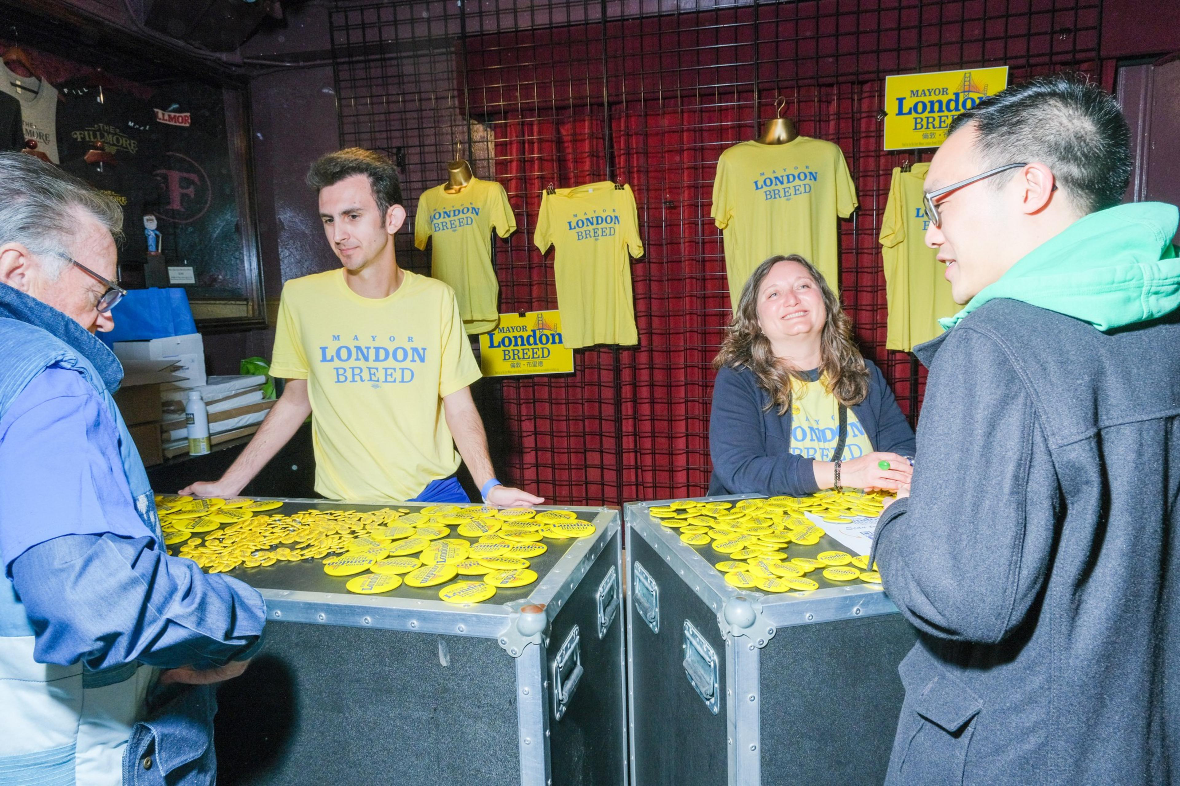 Two people behind a stall handing out yellow stickers and t-shirts with &quot;Mayor London Breed&quot; printed on them to visitors.