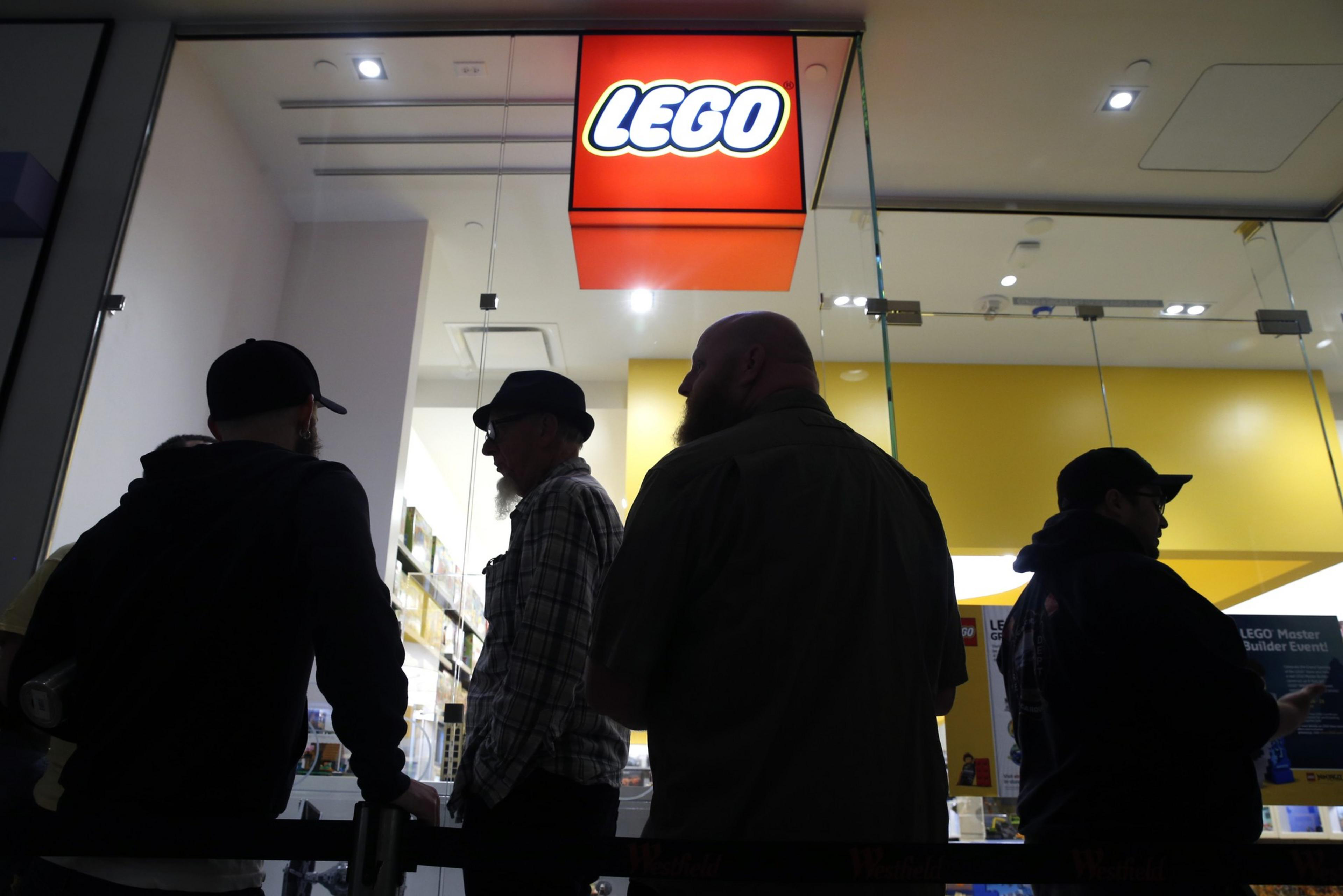 Silhouetted figures stand before a brightly lit LEGO store with a large red and yellow logo.