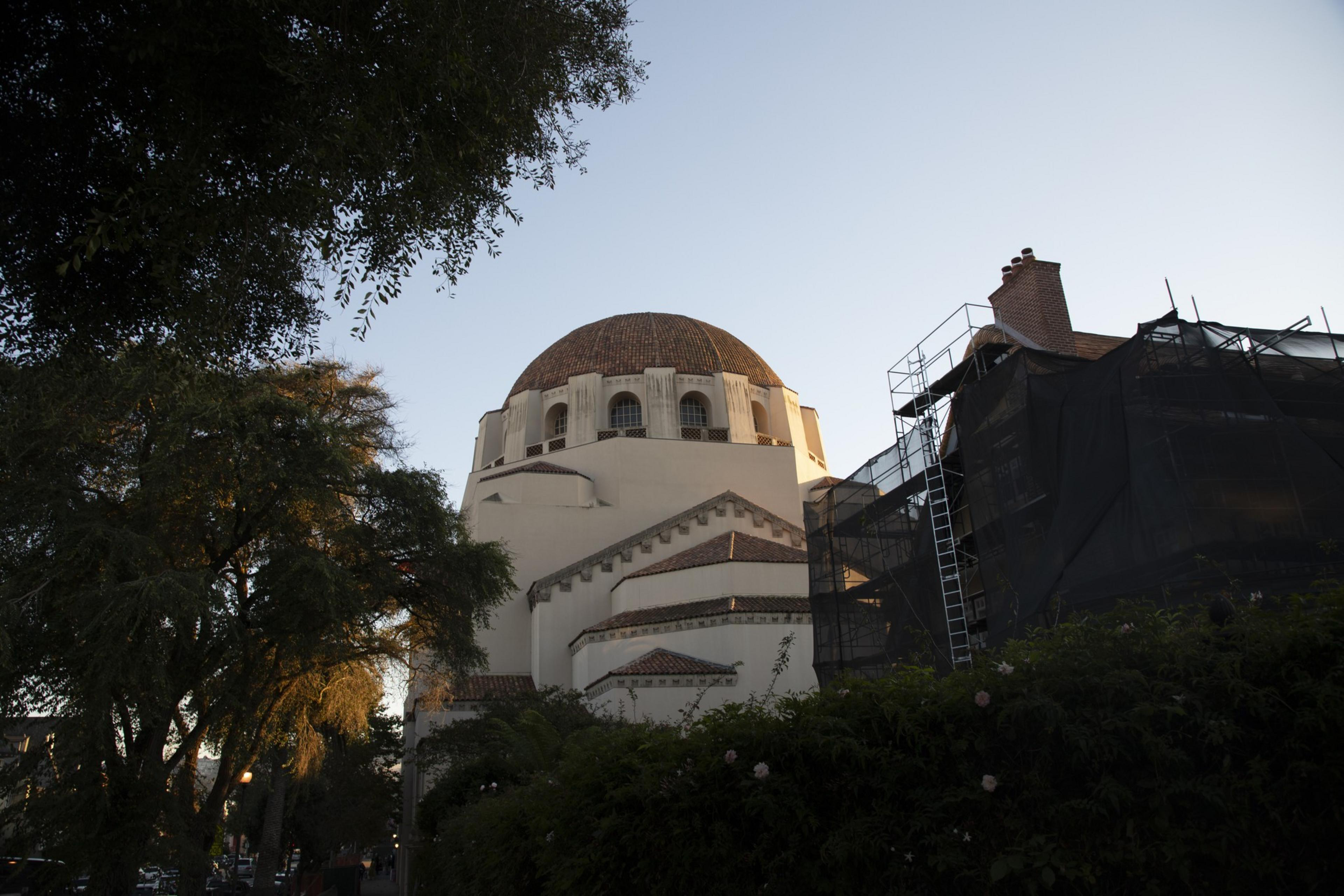 A large domed building with intricate architectural details is partially obscured by trees and scaffolding, set against a clear evening sky.