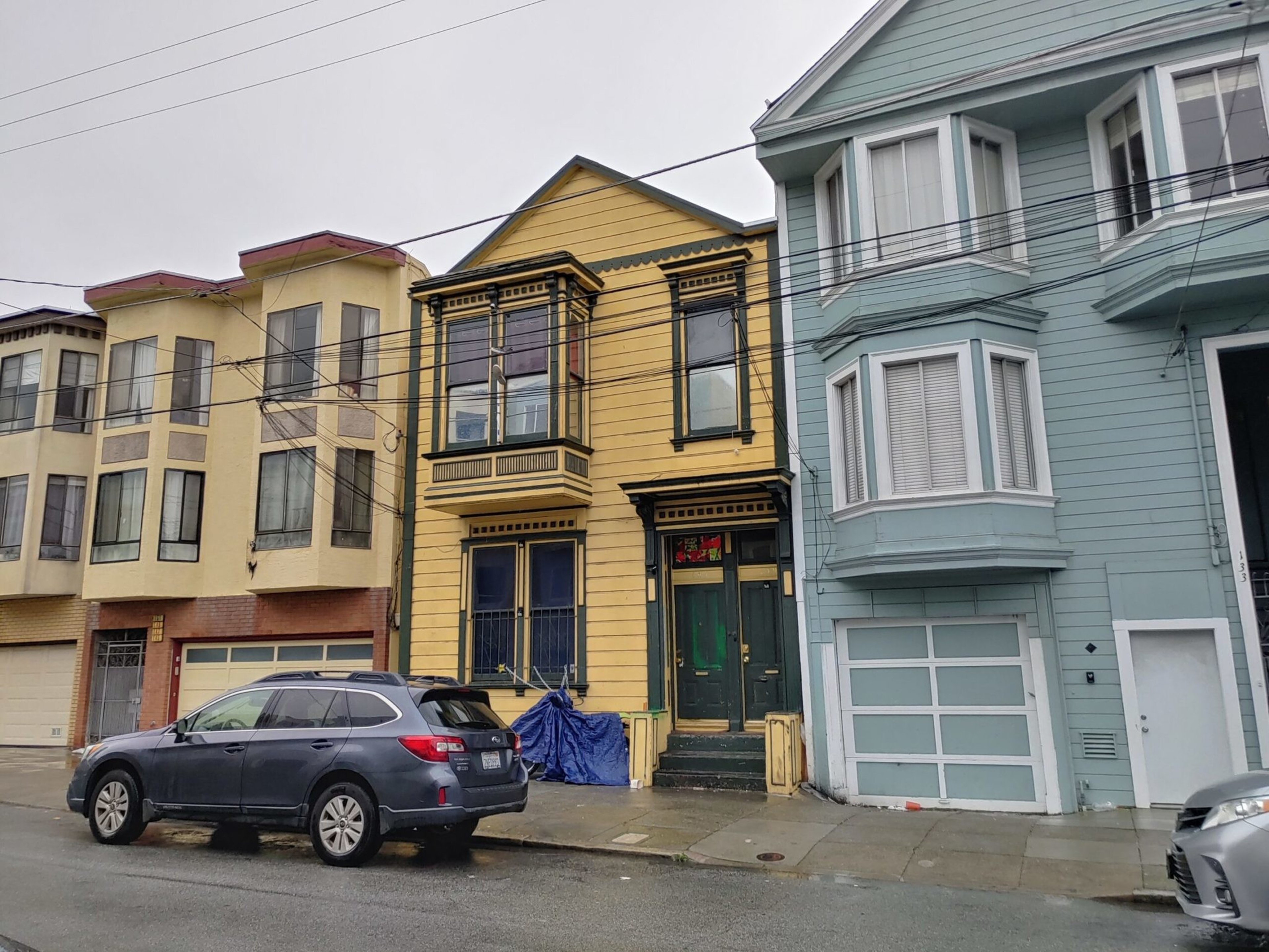 A yellow Victorian-style house sits between beige and light blue houses on a wet street with a dark gray SUV parked in front.