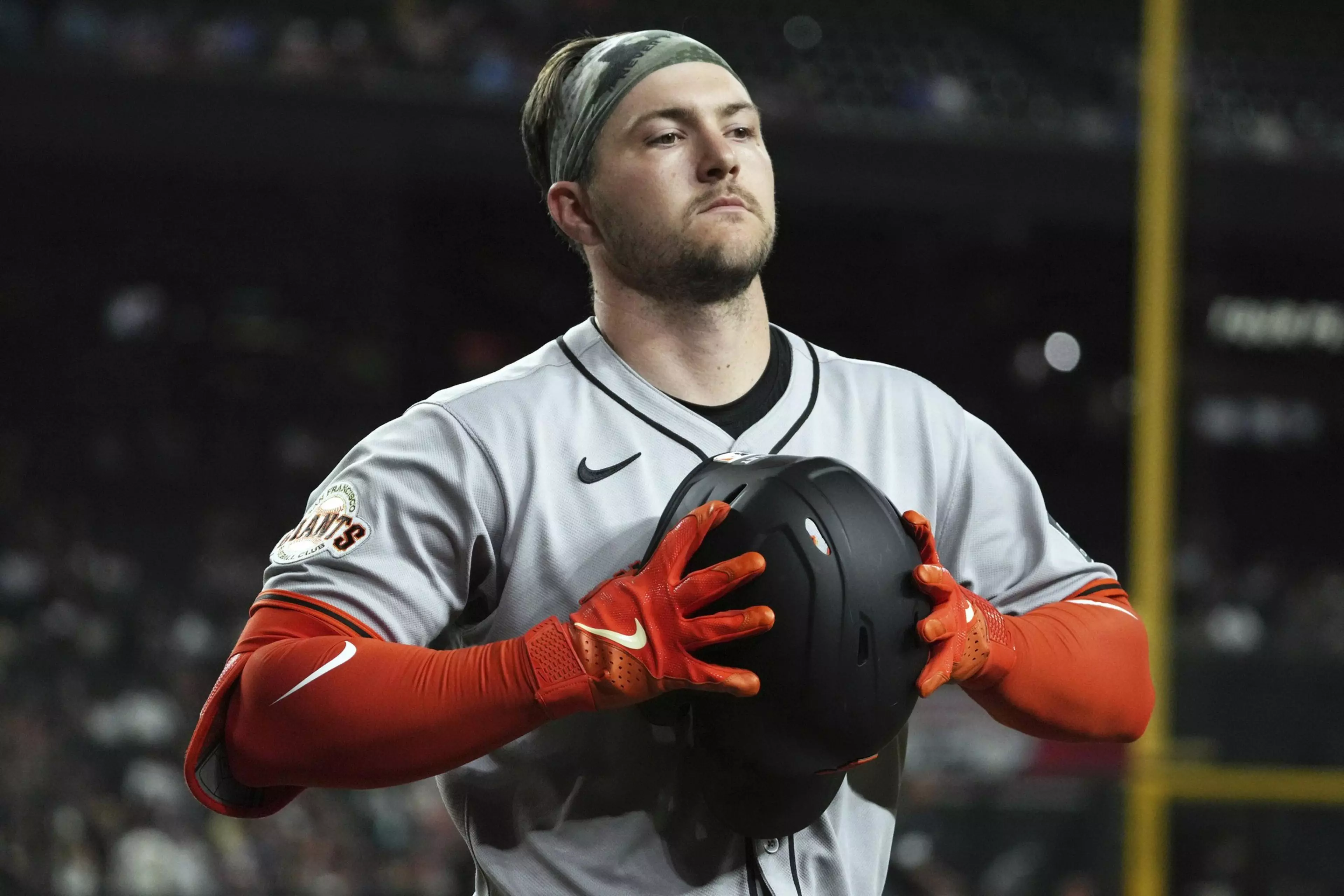 A baseball player in a gray uniform and orange gloves holds a black helmet against his chest. He wears a patterned headband, and the setting appears to be a stadium.