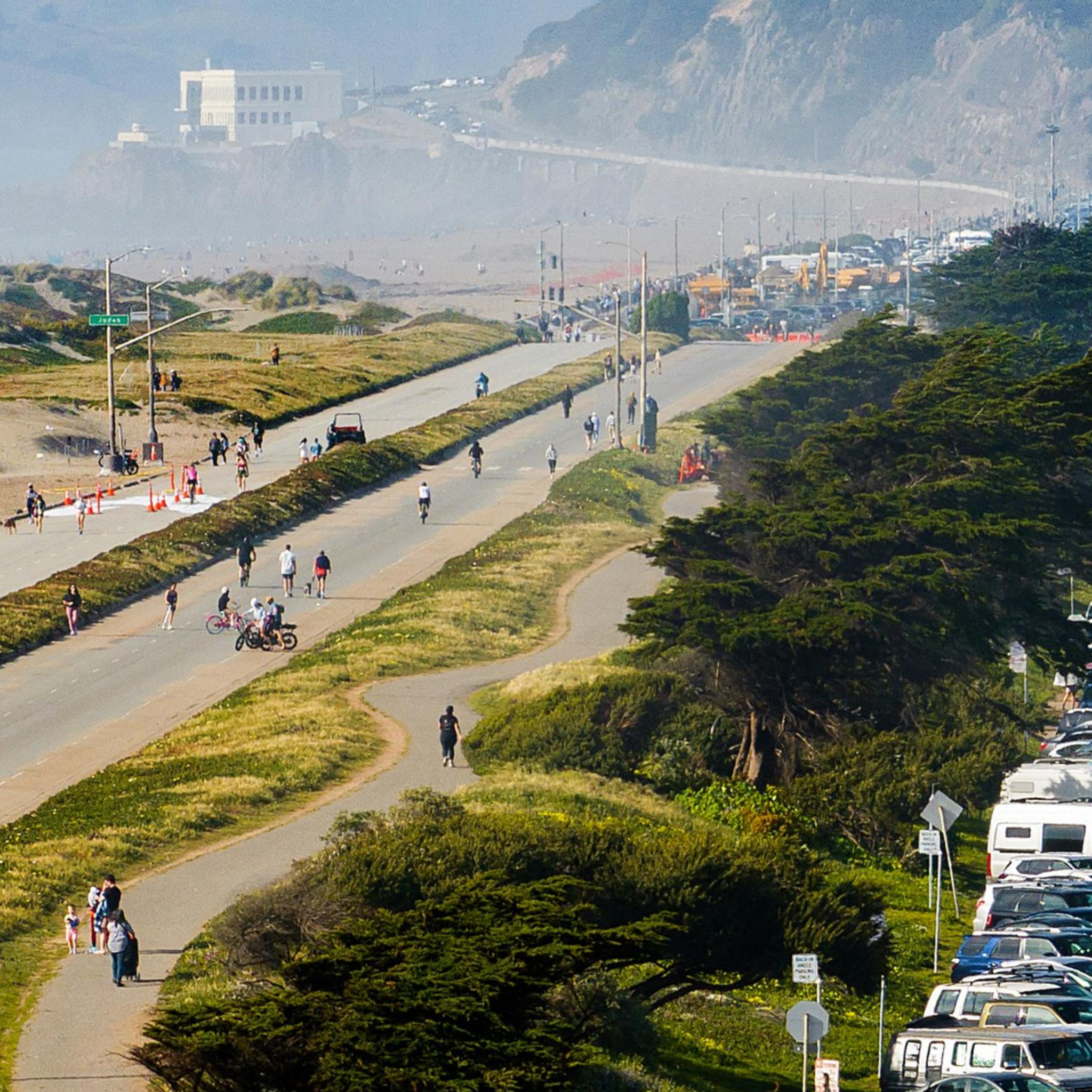 A busy road with heavy traffic and parked cars is adjacent to a separate pedestrian path and bike lane. People are walking and cycling under clear skies.