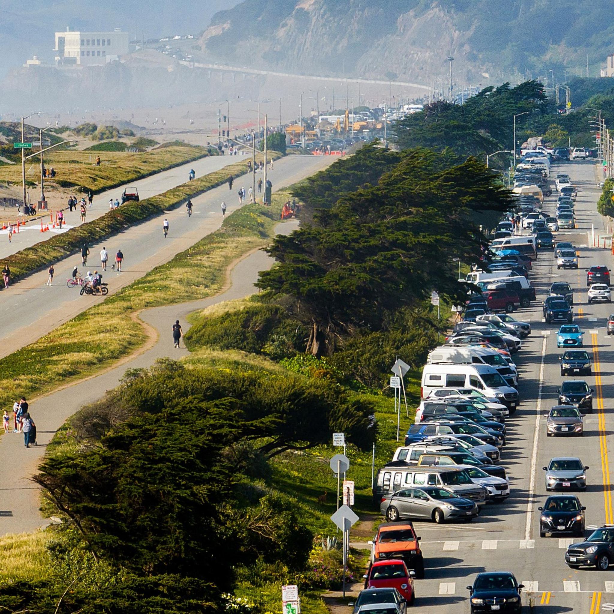 A busy road with heavy traffic and parked cars is adjacent to a separate pedestrian path and bike lane. People are walking and cycling under clear skies.