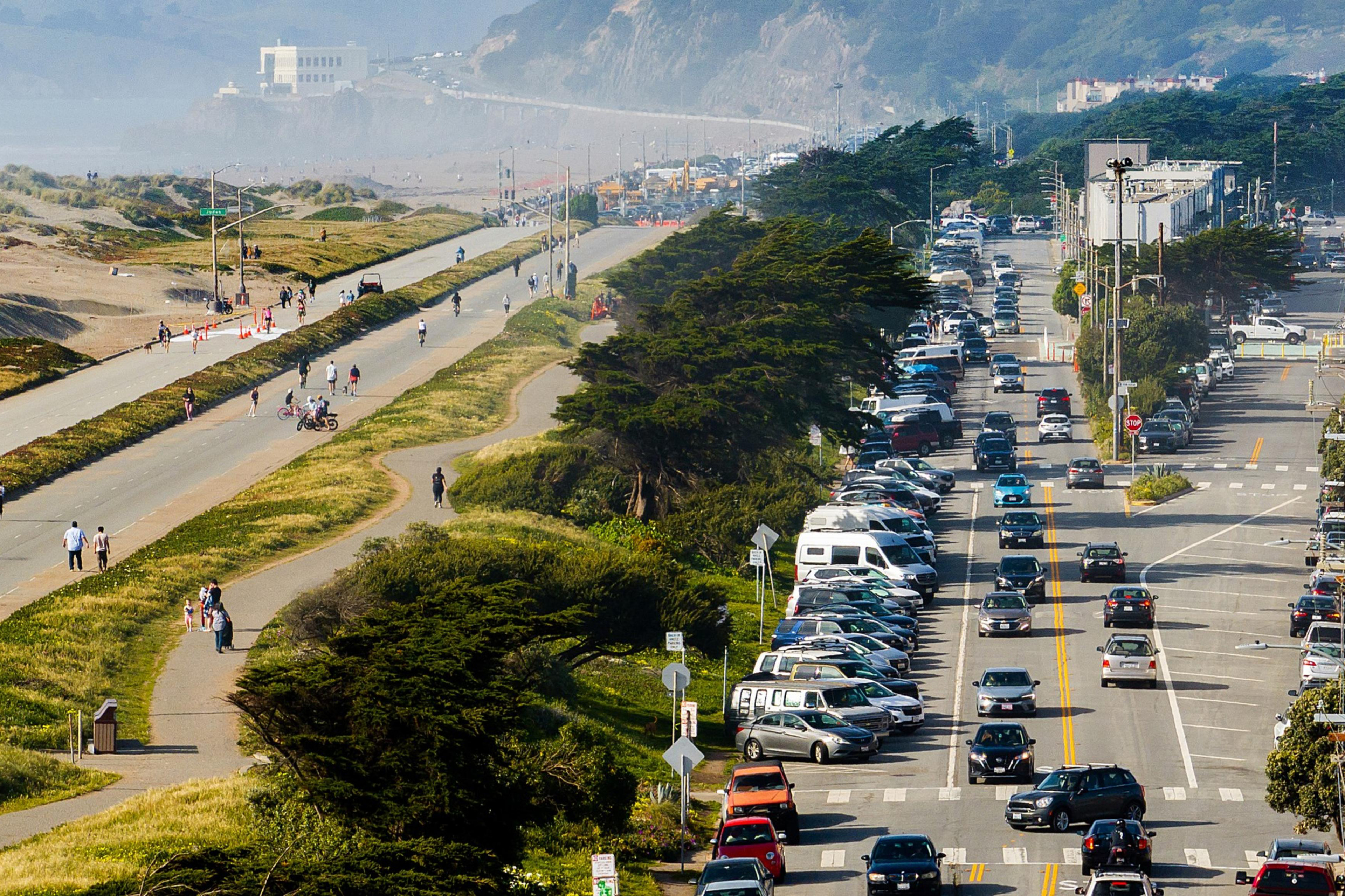 A busy road with heavy traffic and parked cars is adjacent to a separate pedestrian path and bike lane. People are walking and cycling under clear skies.