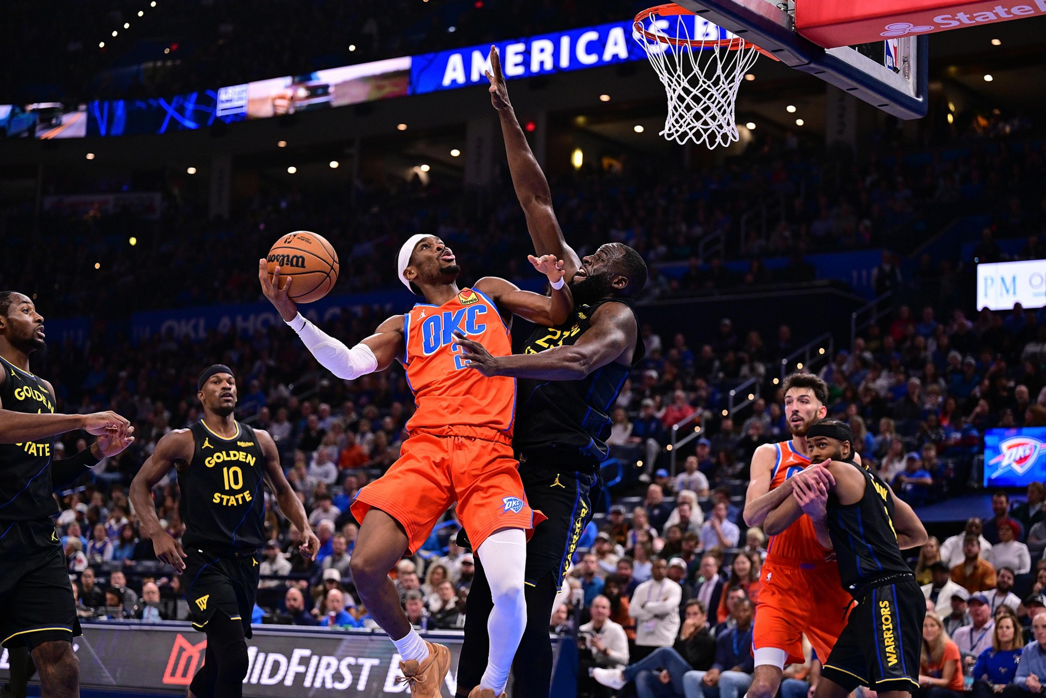 An Oklahoma City player in orange jumps to score while a Golden State defender in black tries to block during a crowded basketball game.