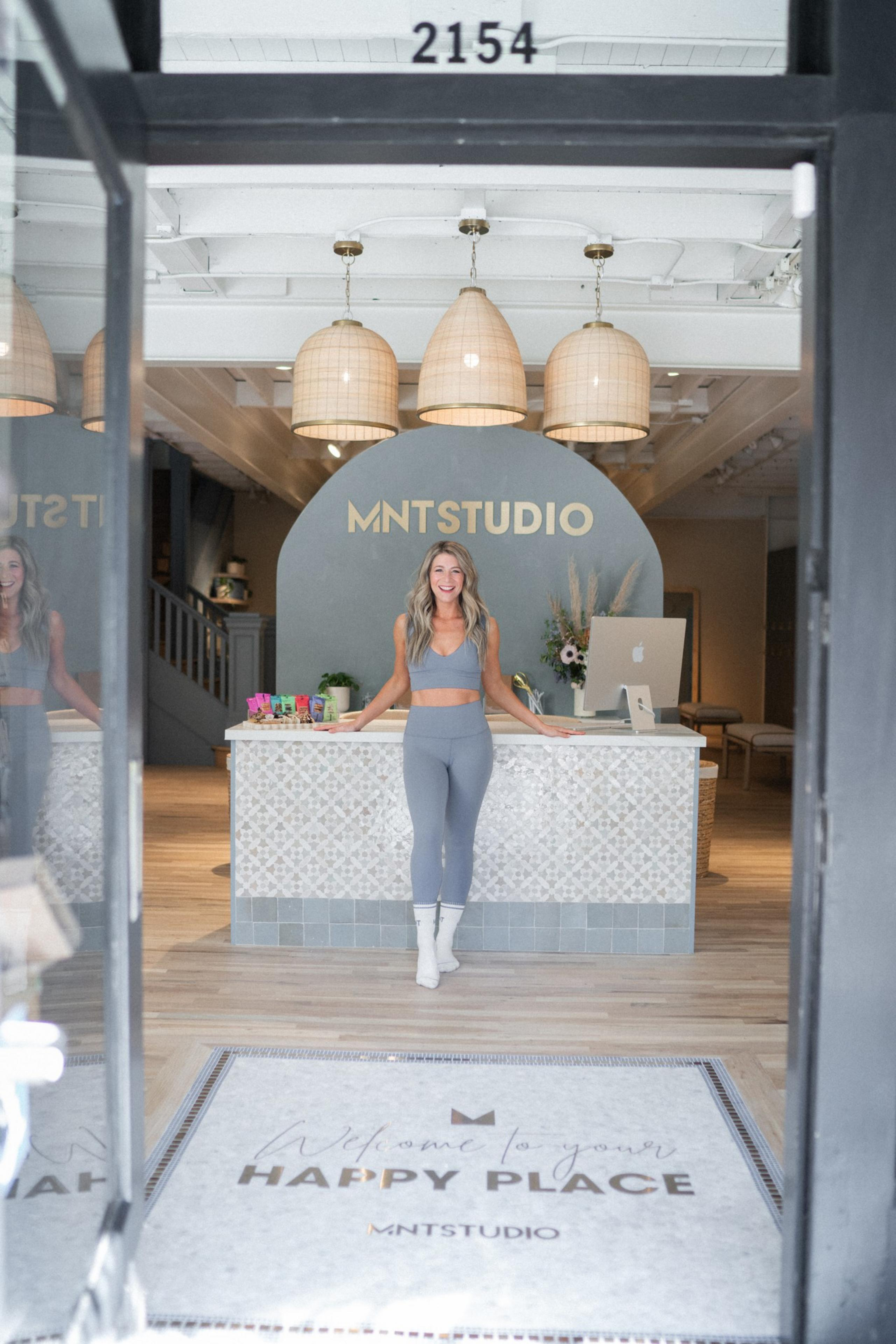 A person in workout attire stands smiling at the entrance of MNT Studio, with a reception desk, decorative lights, flowers, and a mat stating &quot;Welcome to your Happy Place.&quot;