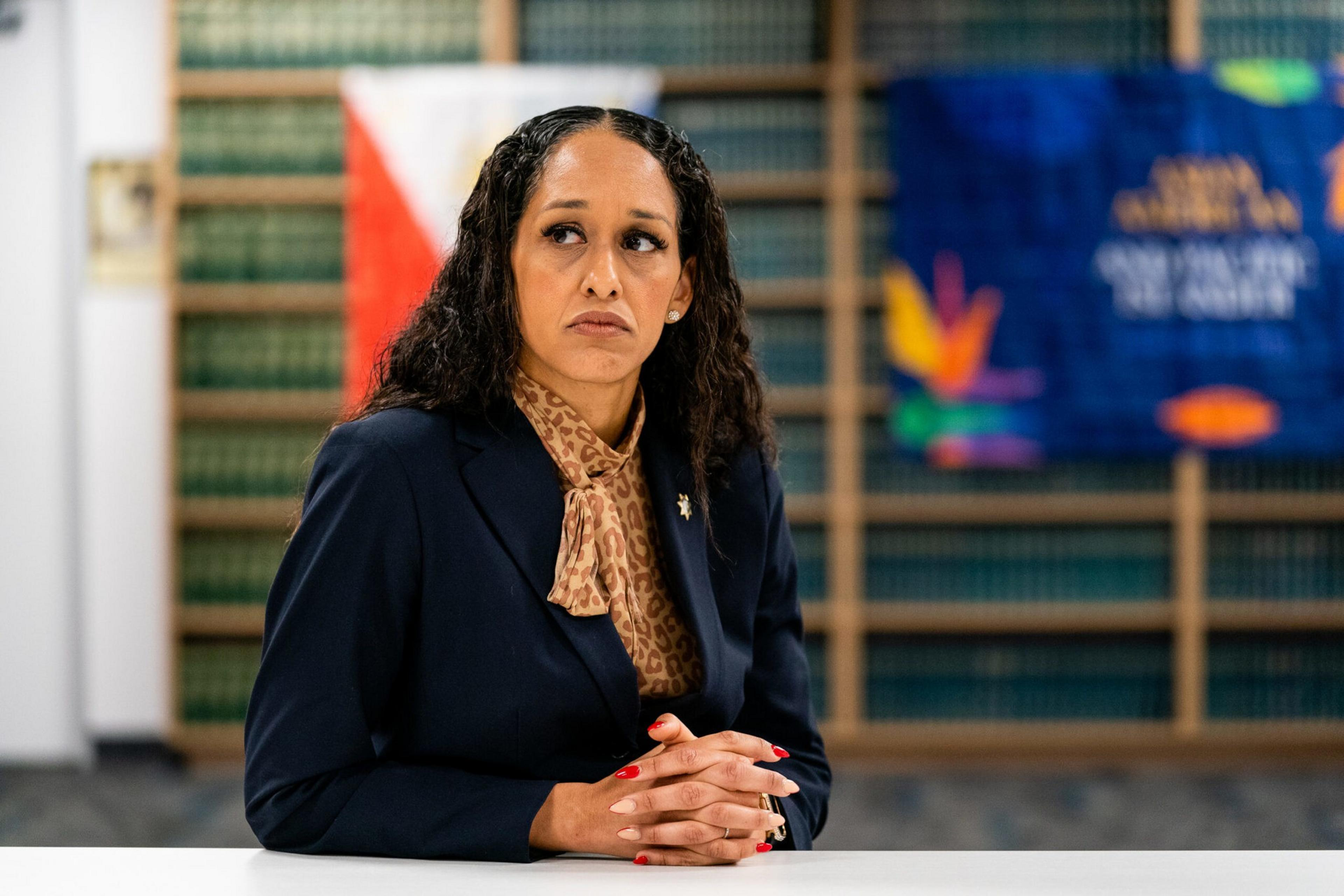 A woman with curly hair wearing a dark blazer and leopard-print scarf sits at a table with hands clasped, looking pensively to the side.