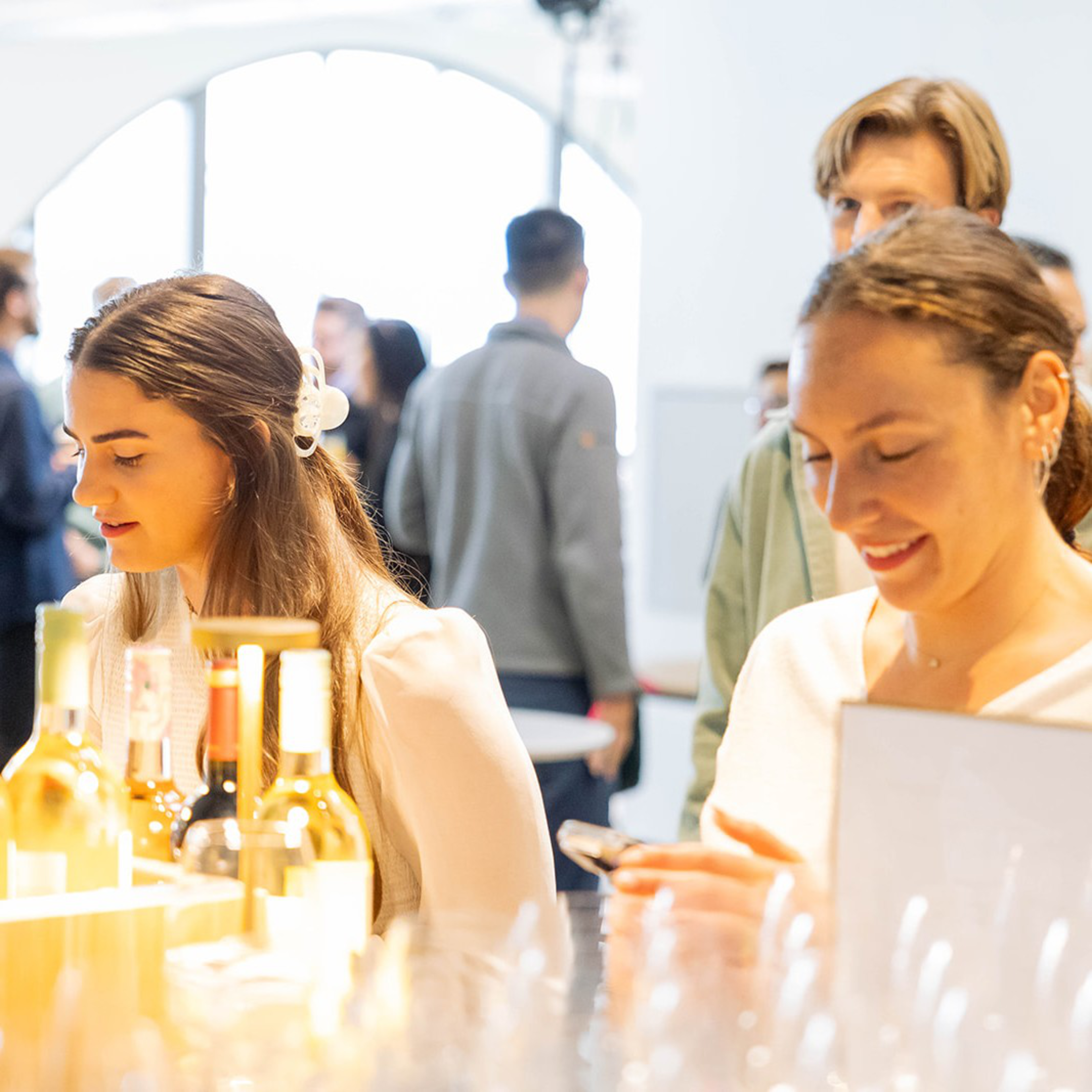 Two women are at a brightly lit bar with wine bottles in front of them. One is looking down, the other is smiling at her phone. People are blurred in the background.
