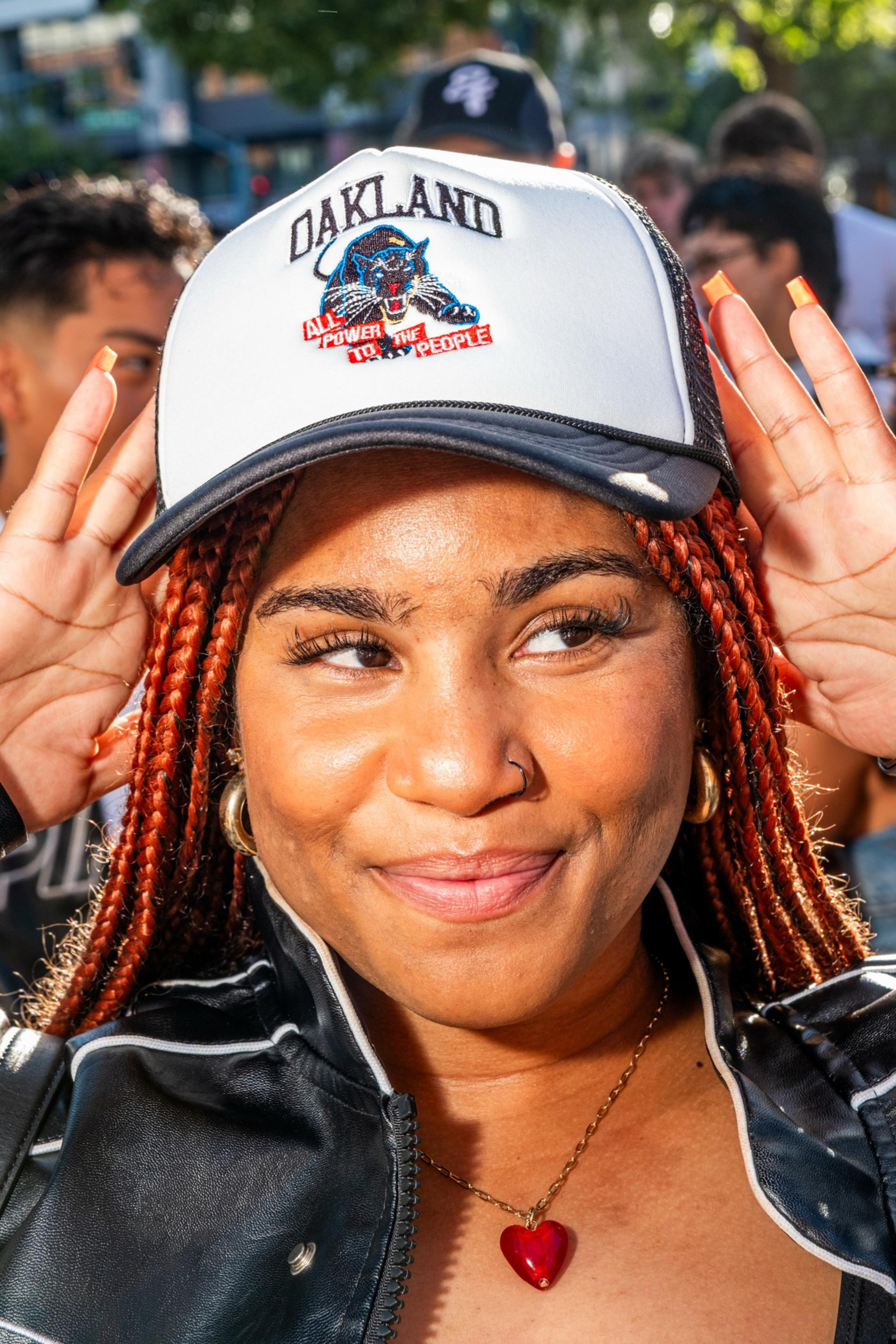 A person with braided hair smiles, wearing a cap with &quot;Oakland&quot; and a panther design. They have gold hoop earrings, a leather jacket, and a red heart necklace.