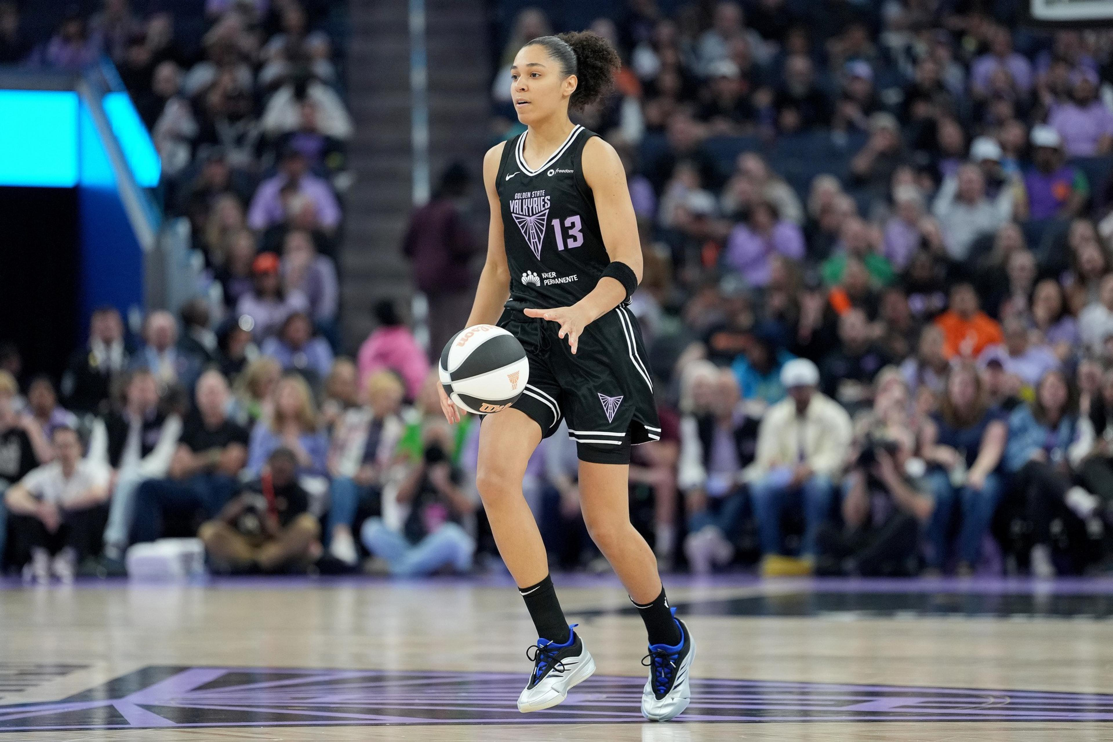 A basketball player in a black uniform dribbles the ball on a court, while a crowd watches from the stands.