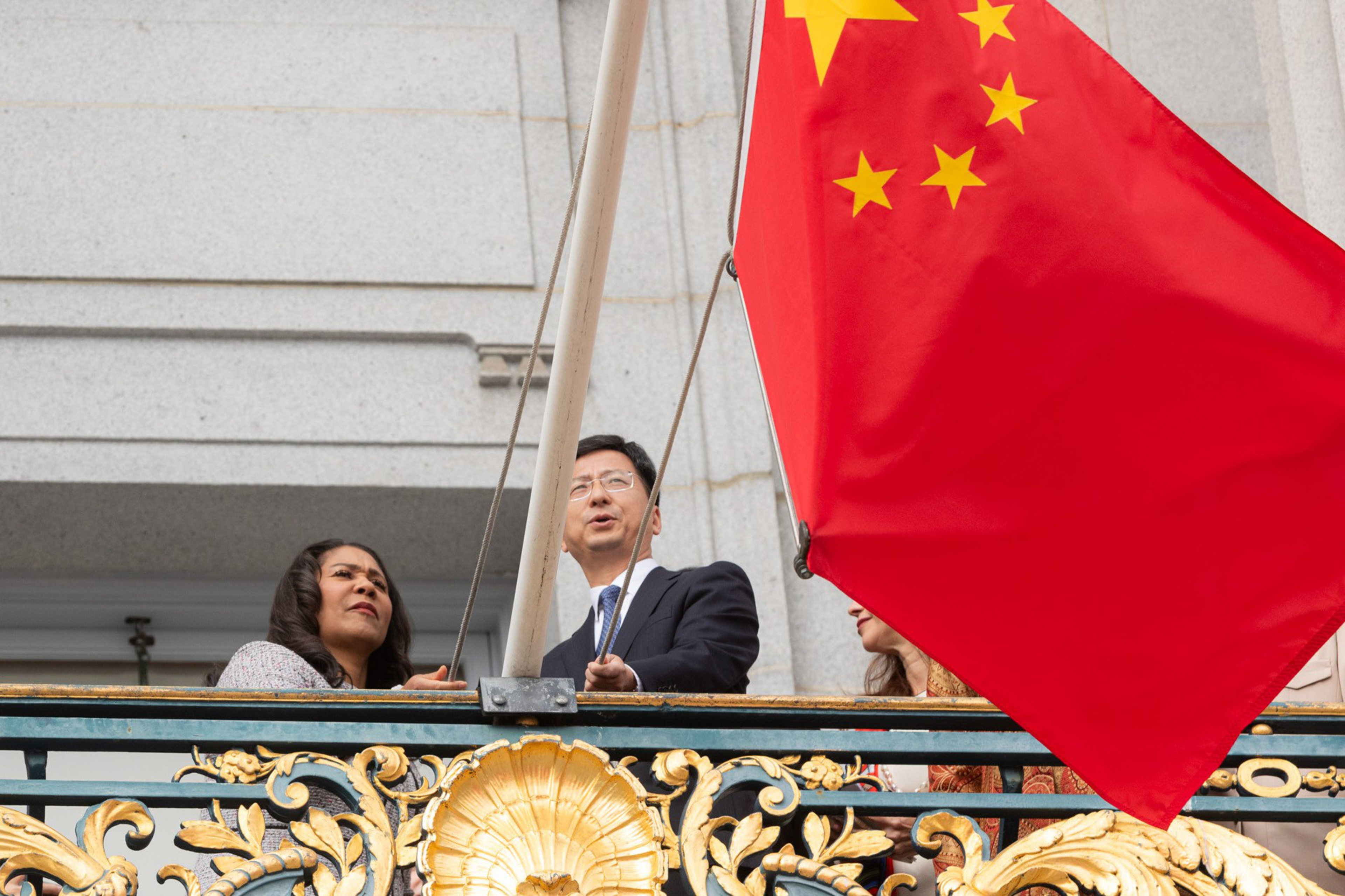 Two people stand behind a Chinese flag on a balcony with a decorative railing.
