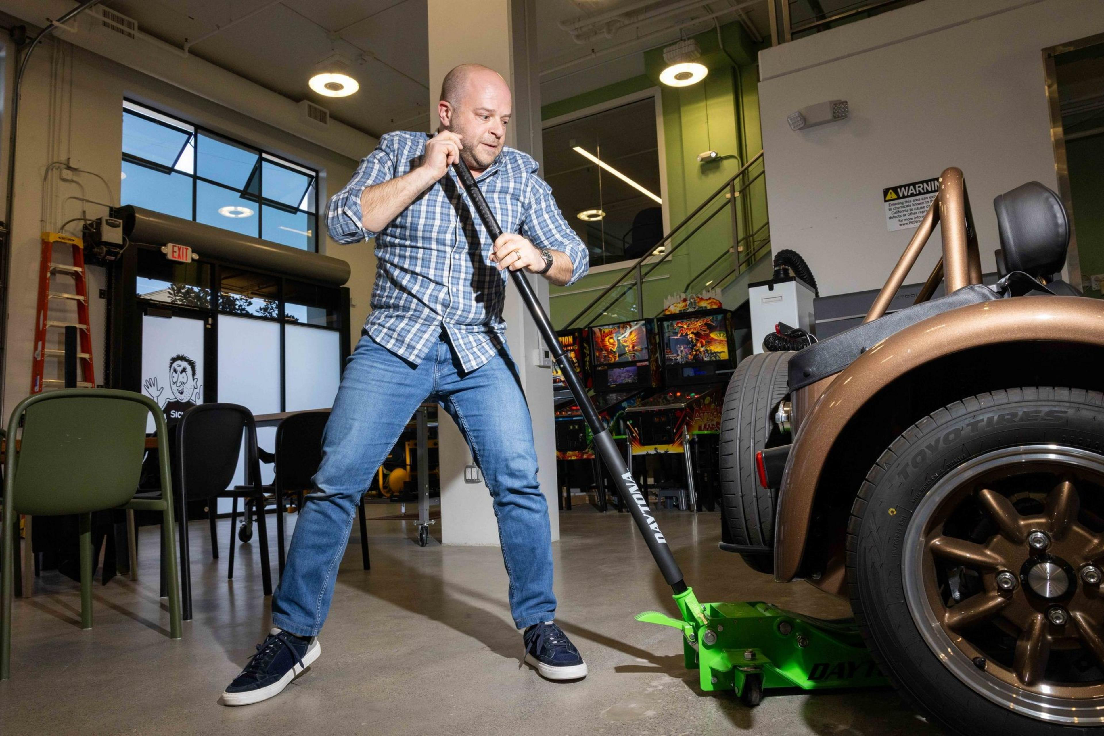 A man in a plaid shirt uses a green jack to lift a car indoors, with arcade games and chairs in the background, under bright lighting.