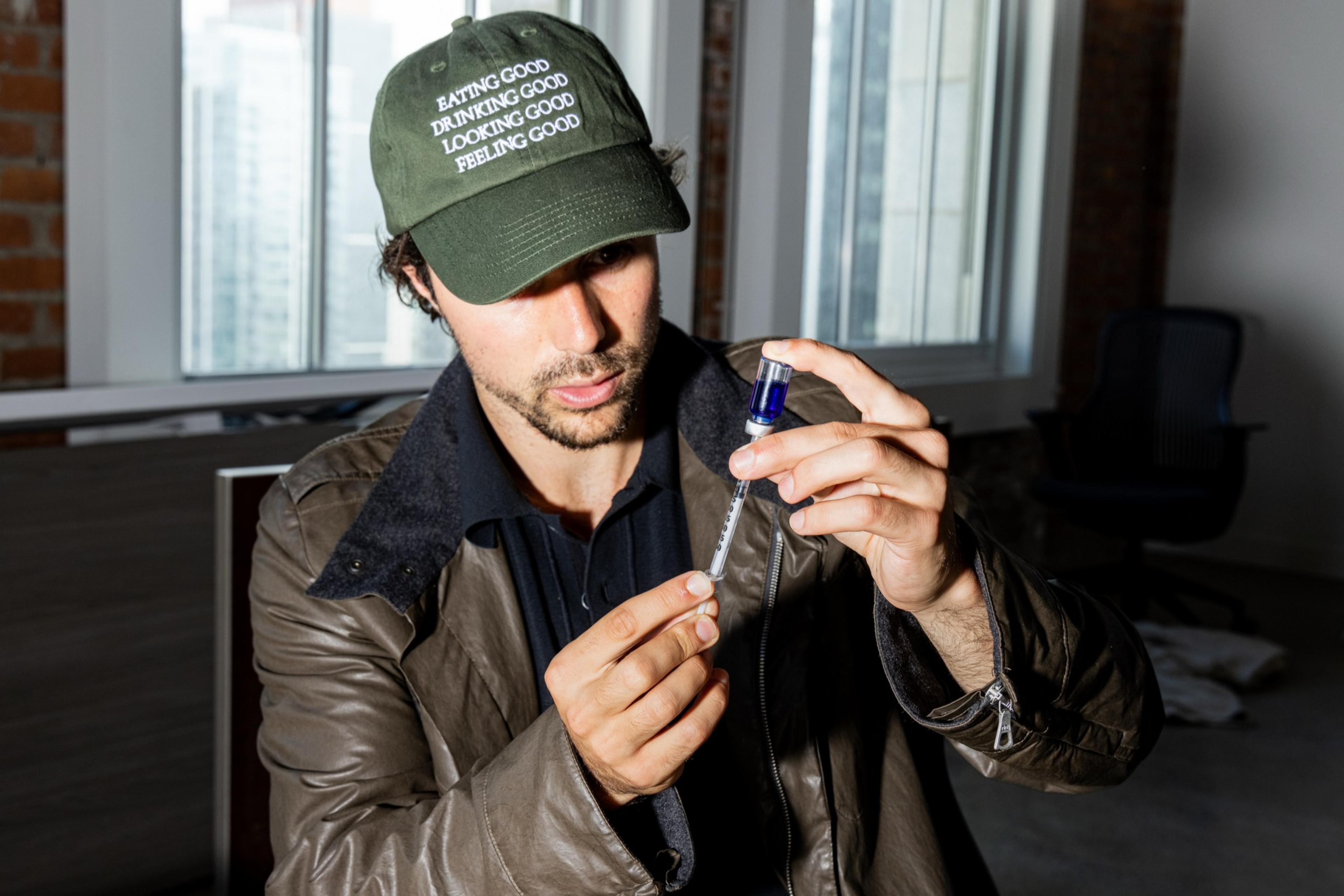 A man wearing a green cap and brown jacket carefully holds a syringe and vial, preparing an injection indoors near large windows.