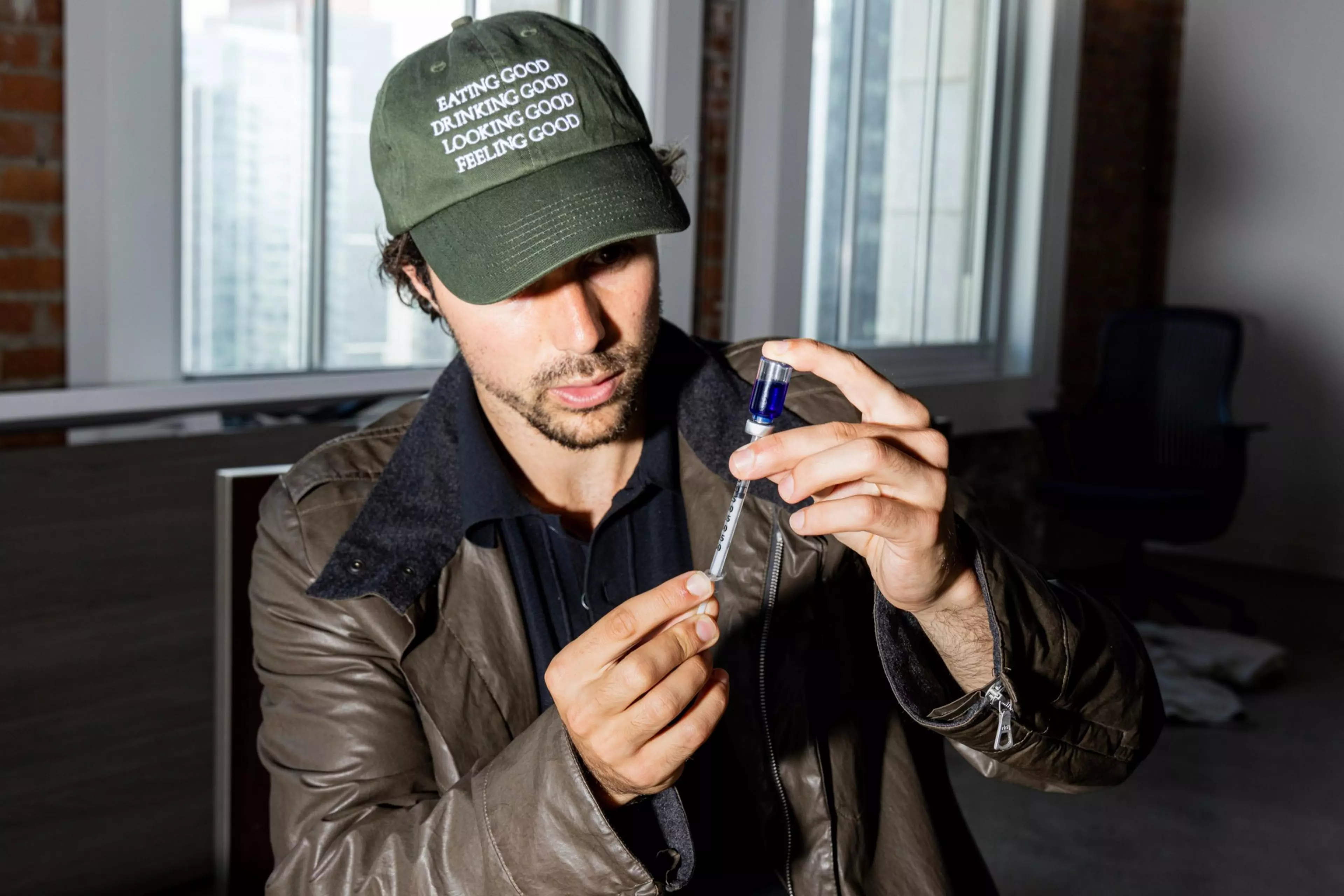 A man wearing a green cap and brown jacket carefully holds a syringe and vial, preparing an injection indoors near large windows.