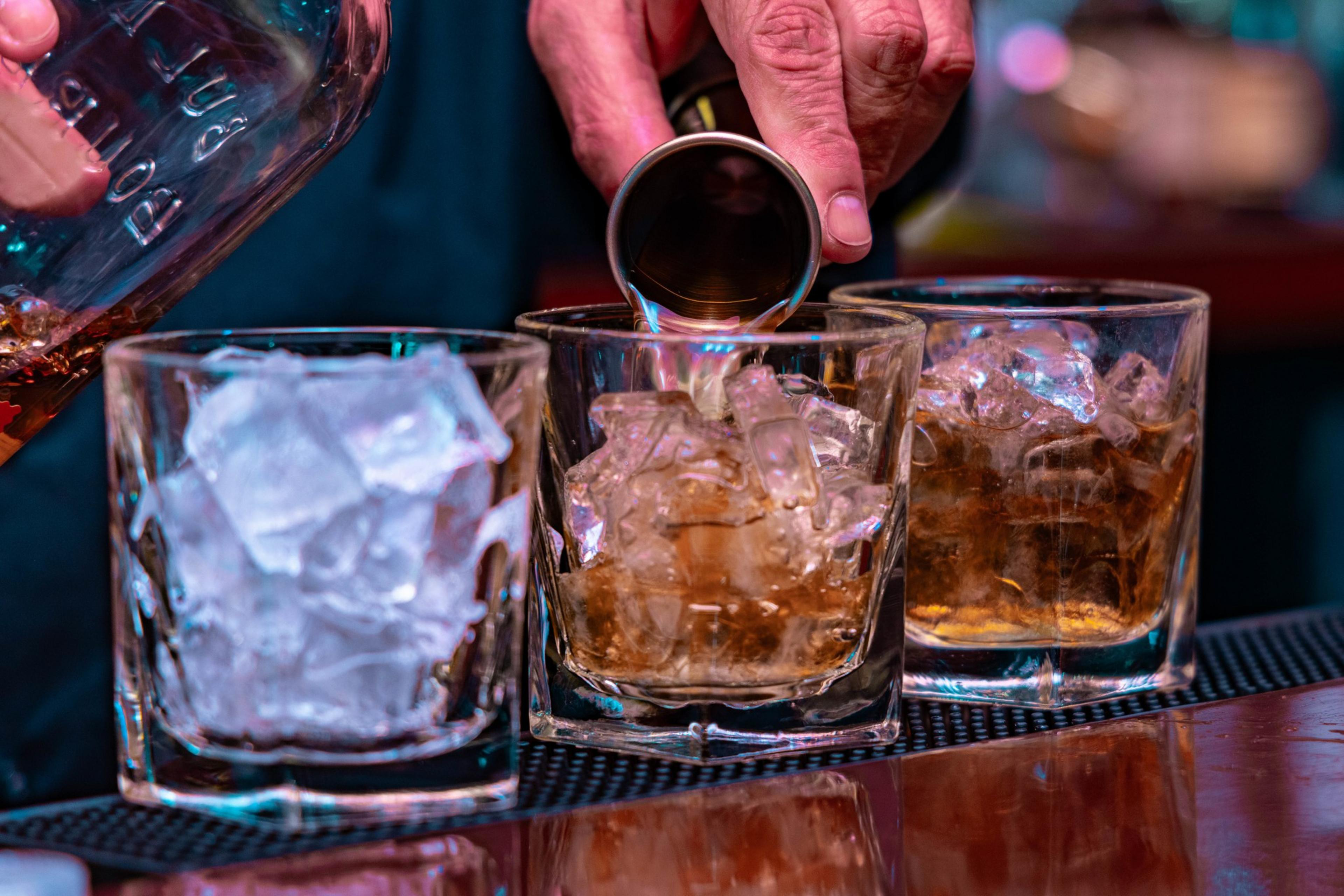 A hand pours alcohol into one of three glasses atop a polished wooden bar tabletop.