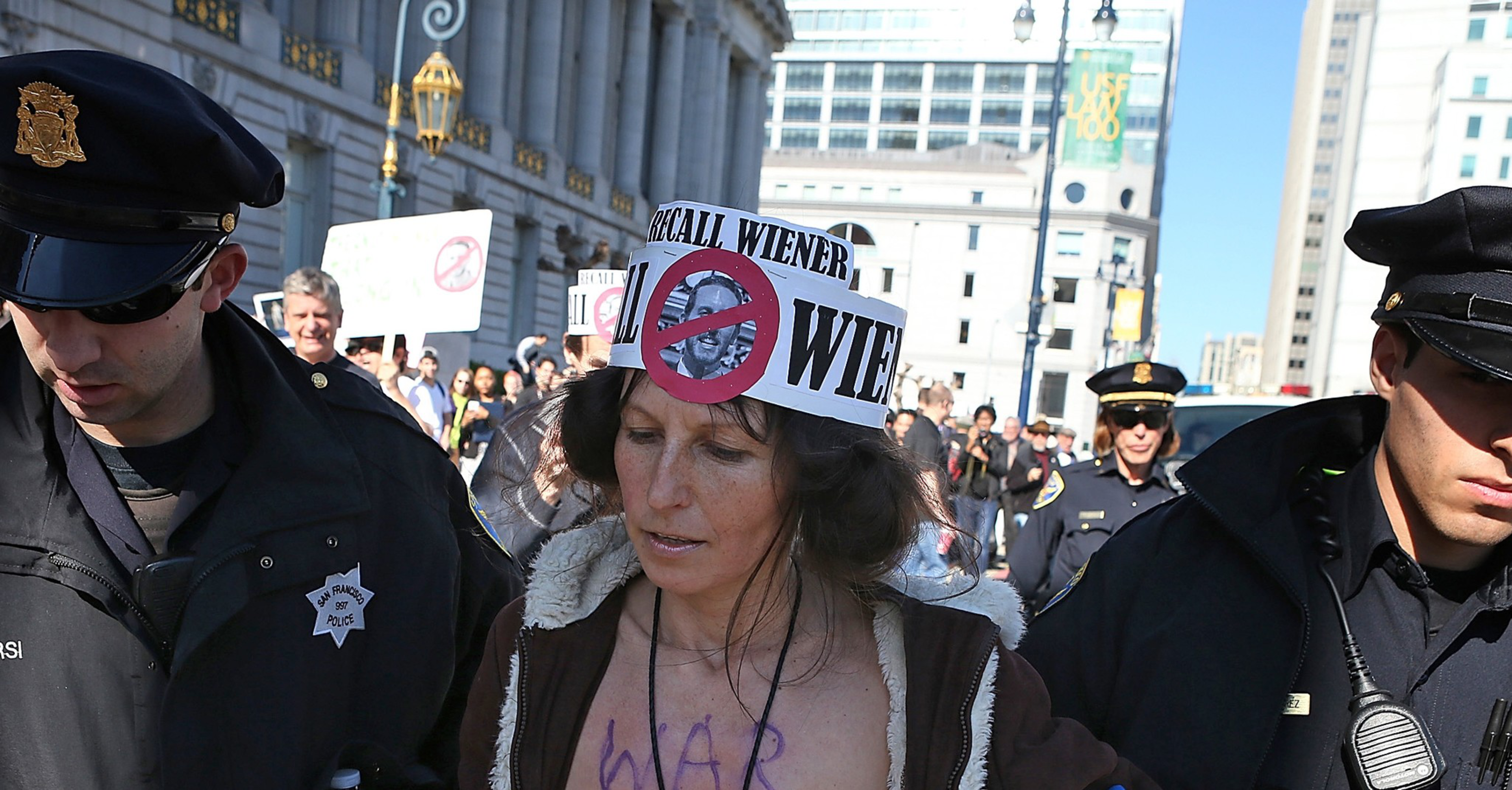 A woman wearing a "Recall Wiener" hat with a crossed-out face is escorted by police officers in a city protest.