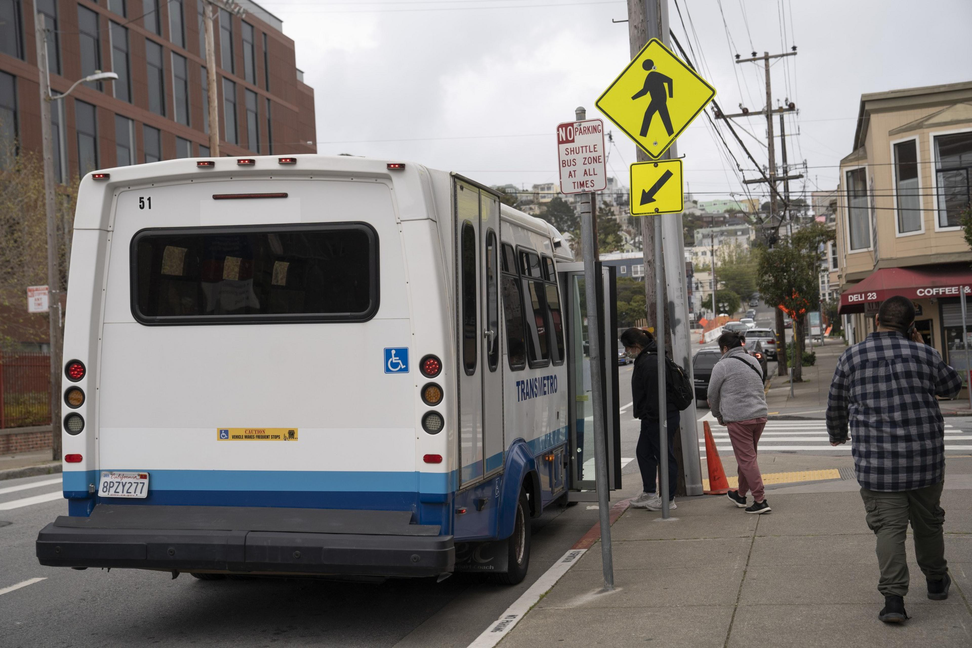 A small white shuttle bus with &quot;Transmetro&quot; is parked on a street. People are boarding it near a crosswalk and a yellow pedestrian sign.