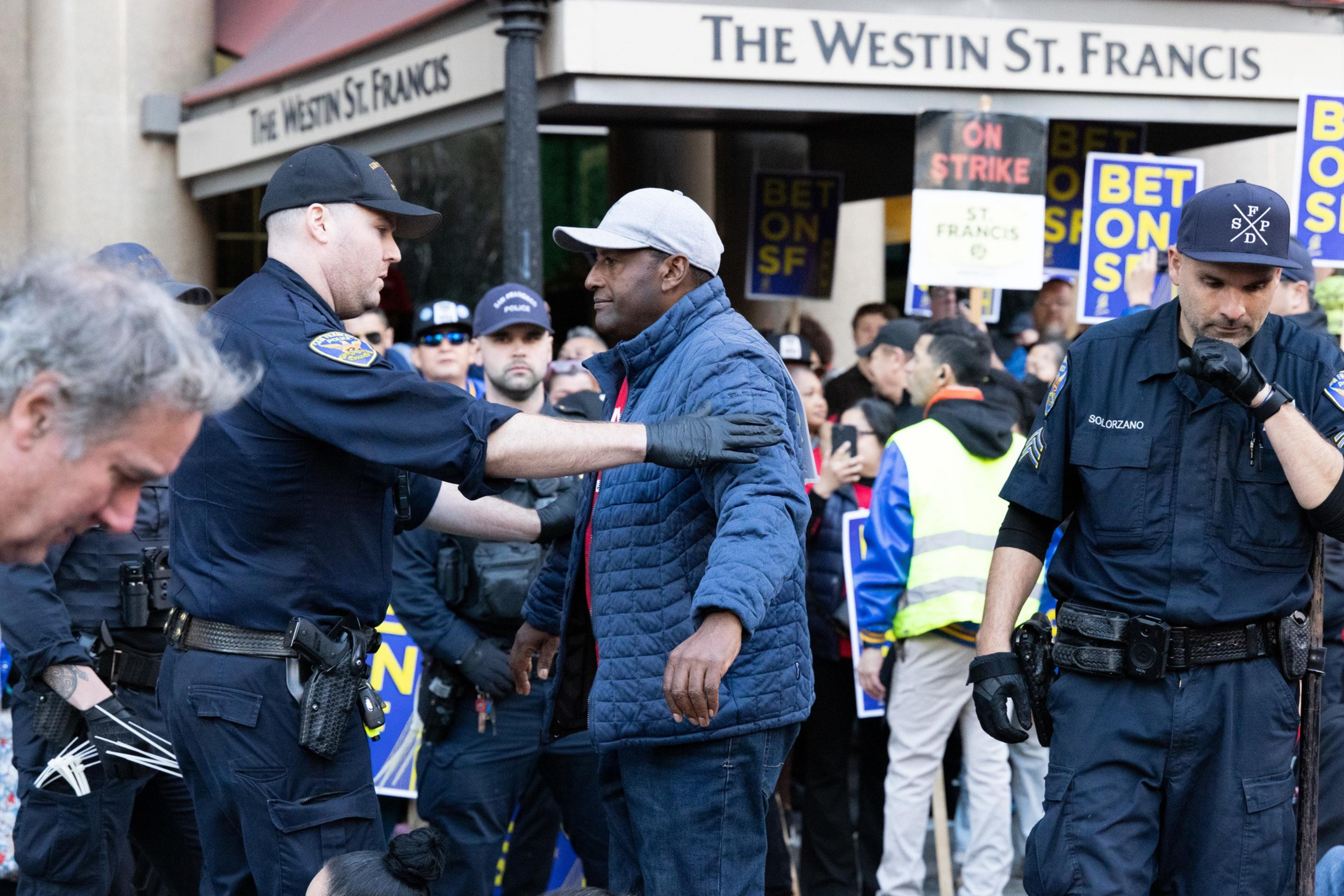 A police officer is interacting with a man in a crowd outside The Westin St. Francis hotel. Protesters hold signs reading &quot;BET ON SF&quot; and &quot;ON STRIKE&quot;.