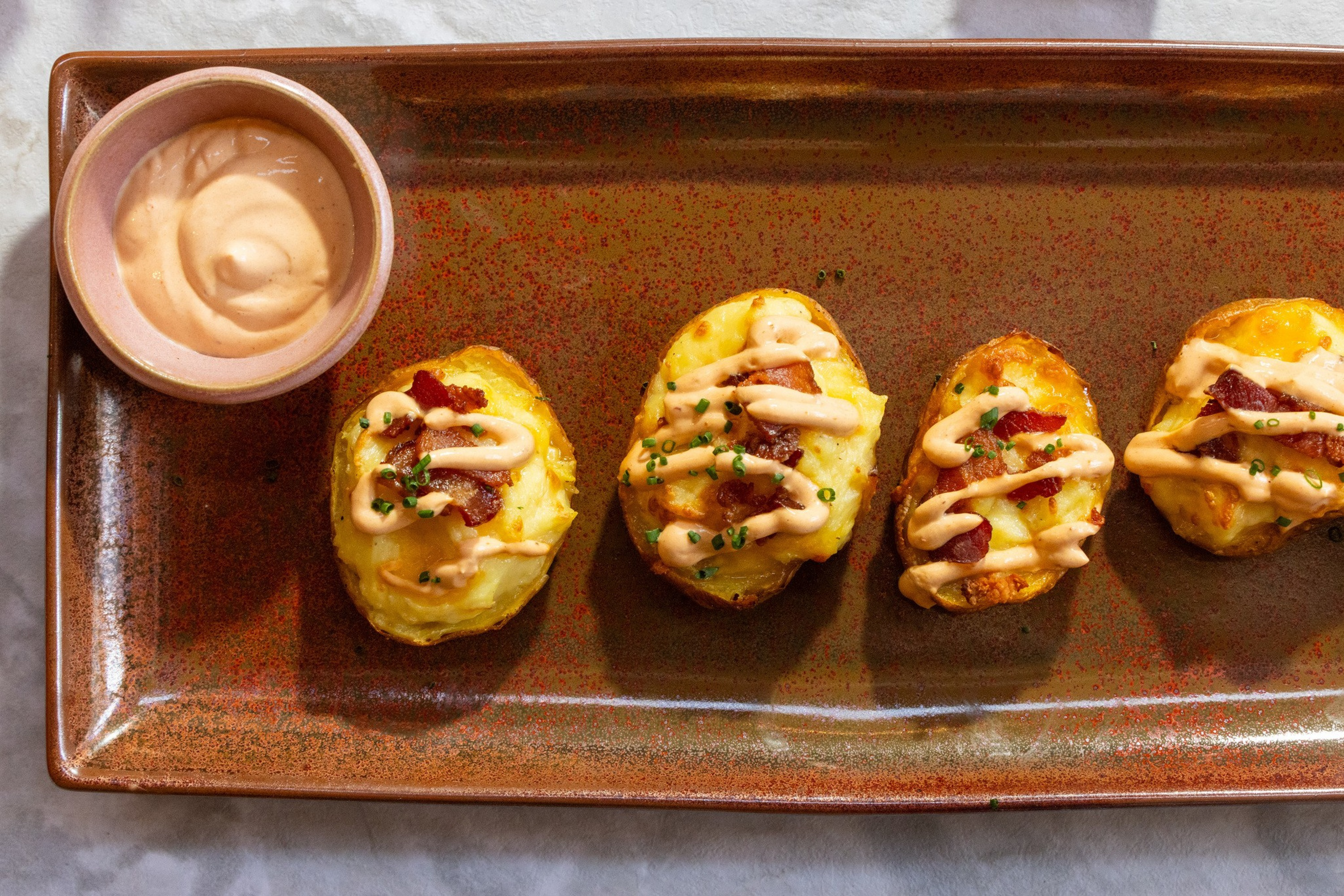 Four twice-baked potato skins topped with bacon bits, chives, and a drizzle of creamy sauce sit on a brown rectangular plate with extra sauce on the side.