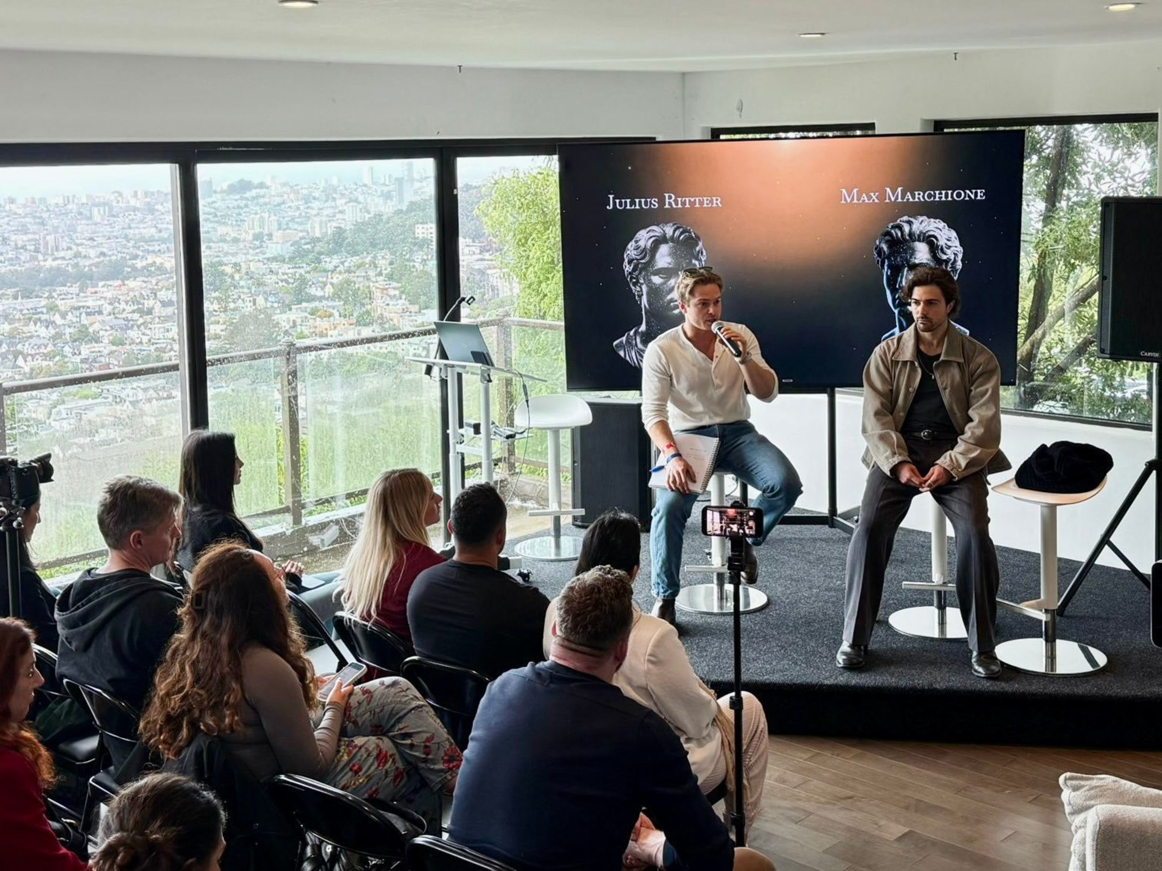 Two men are seated on stage in front of an audience, one speaking into a microphone, with a city view behind and a screen displaying “Julius Ritter” and “Max Marchione.”