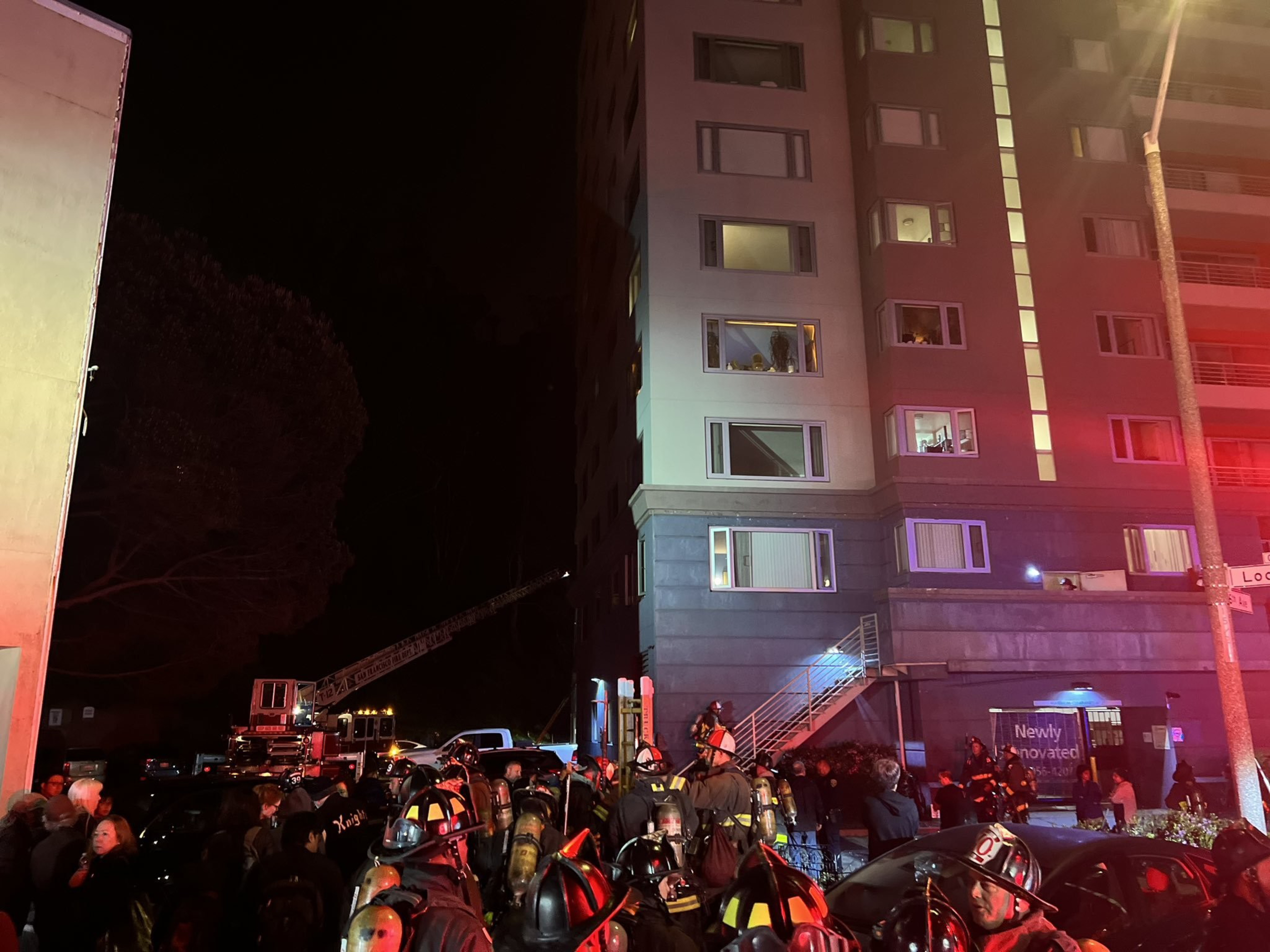 Firefighters in full gear gather outside a multi-story building at night, with a fire truck ladder extended and emergency lights reflecting on the scene.