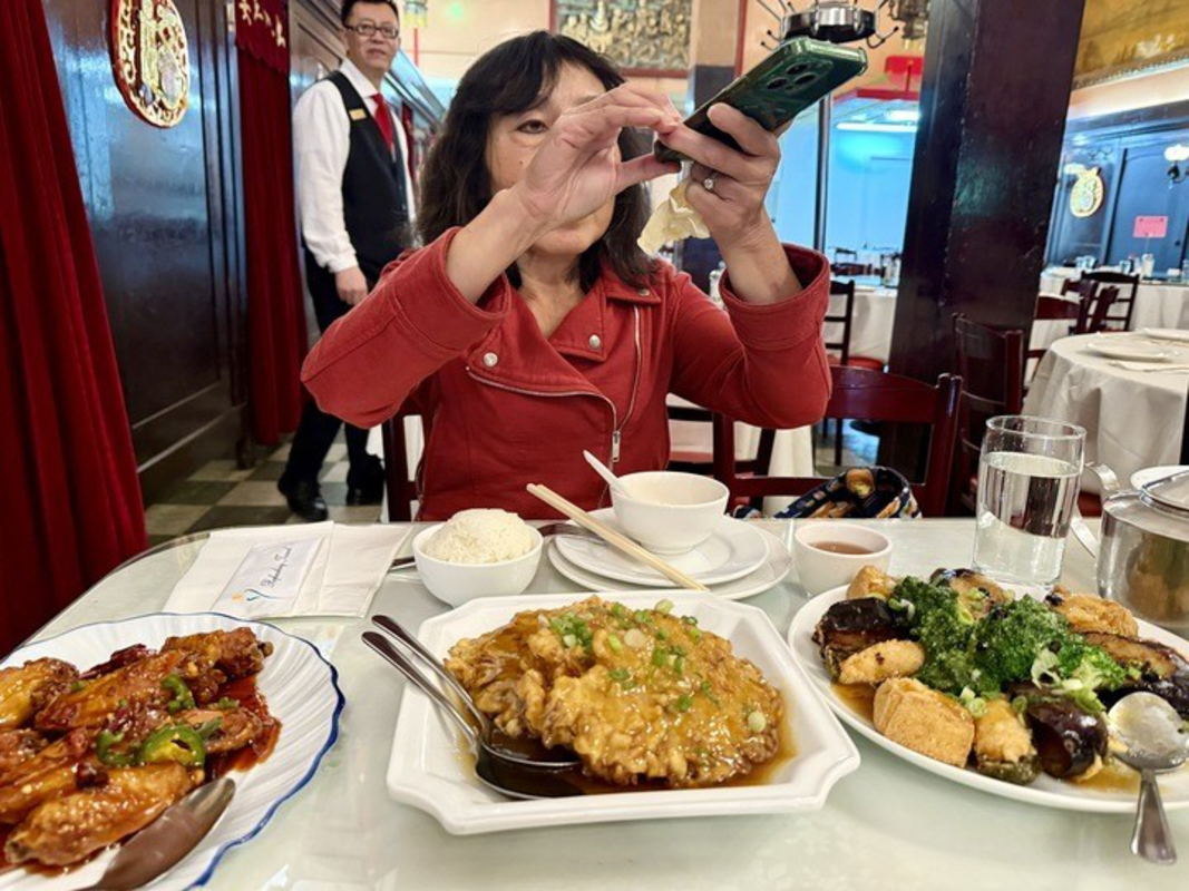 A woman in a red jacket takes a photo of a table filled with various Asian dishes including rice, sauced chicken wings, an egg dish, and a vegetable stir-fry.