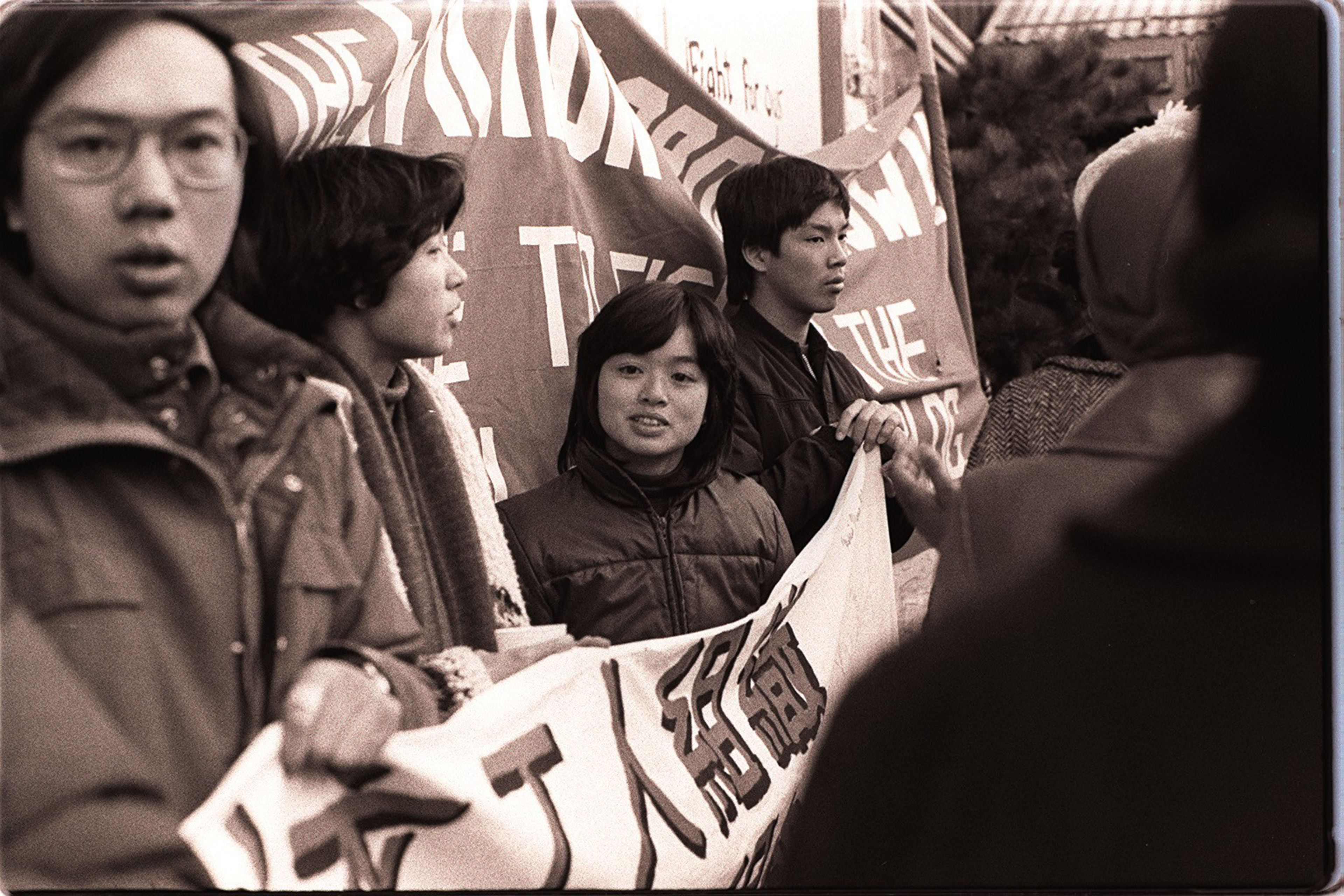 A group of young people hold banners with text, standing close together, appearing to participate in a protest or demonstration outdoors.