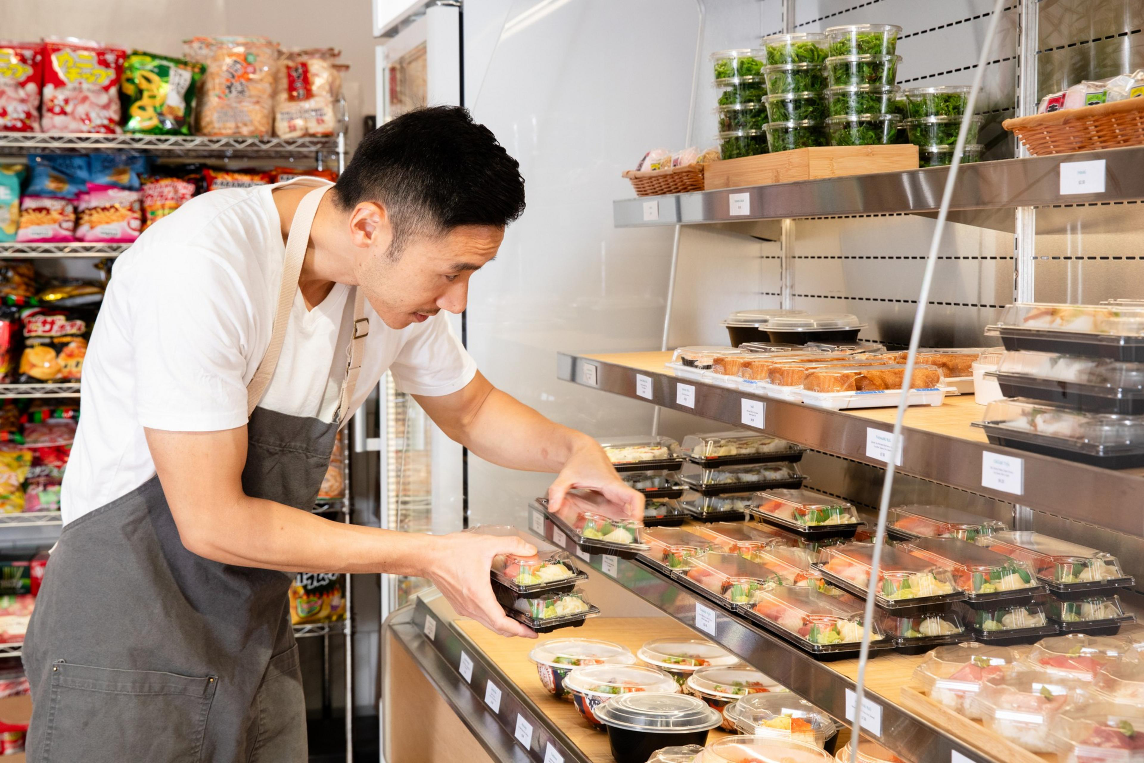 A man in an apron stocks shelves with packaged food in a store. The shelves are filled with pre-made meals and snacks, while various products are displayed behind him.